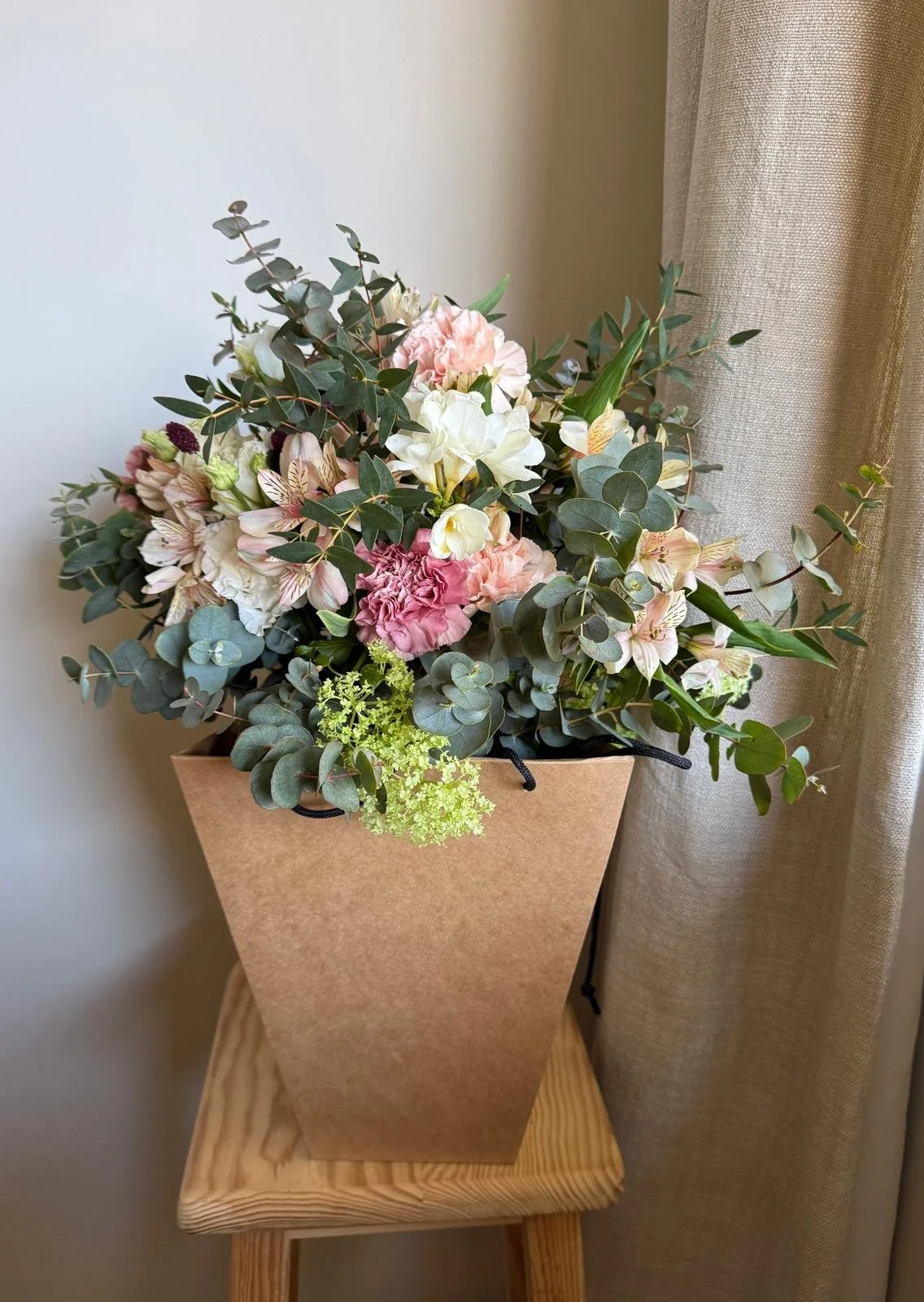 A bouquet of pink and white flowers with green foliage in a brown paper wrapper, placed on a wooden table next to beige curtains.