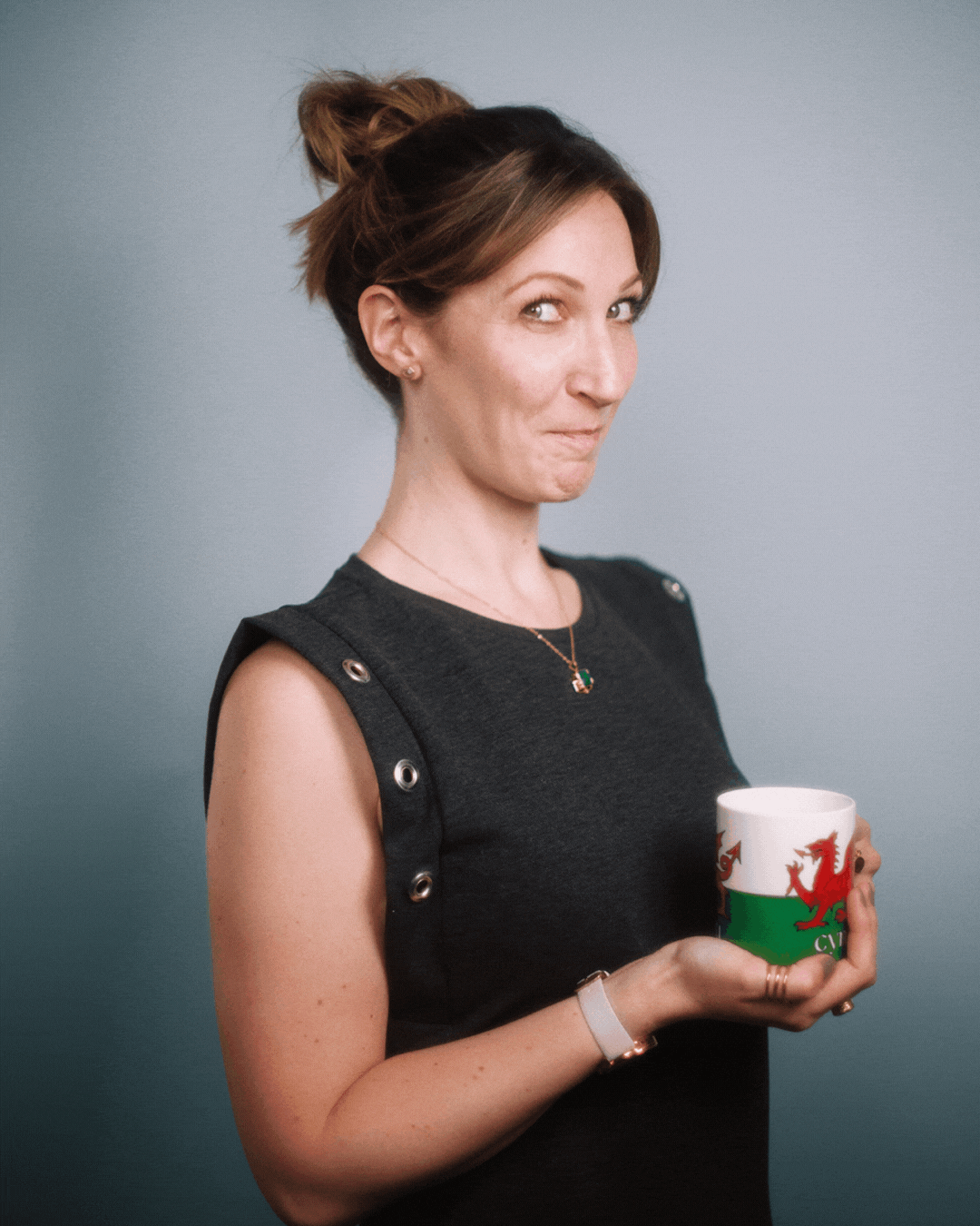 Jess Nesling with short brown hair in a messy bun, wearing a black sleeveless top with metallic button details on the shoulders, holding a white mug with a Welsh dragon emblem, standing against a plain light blue background.