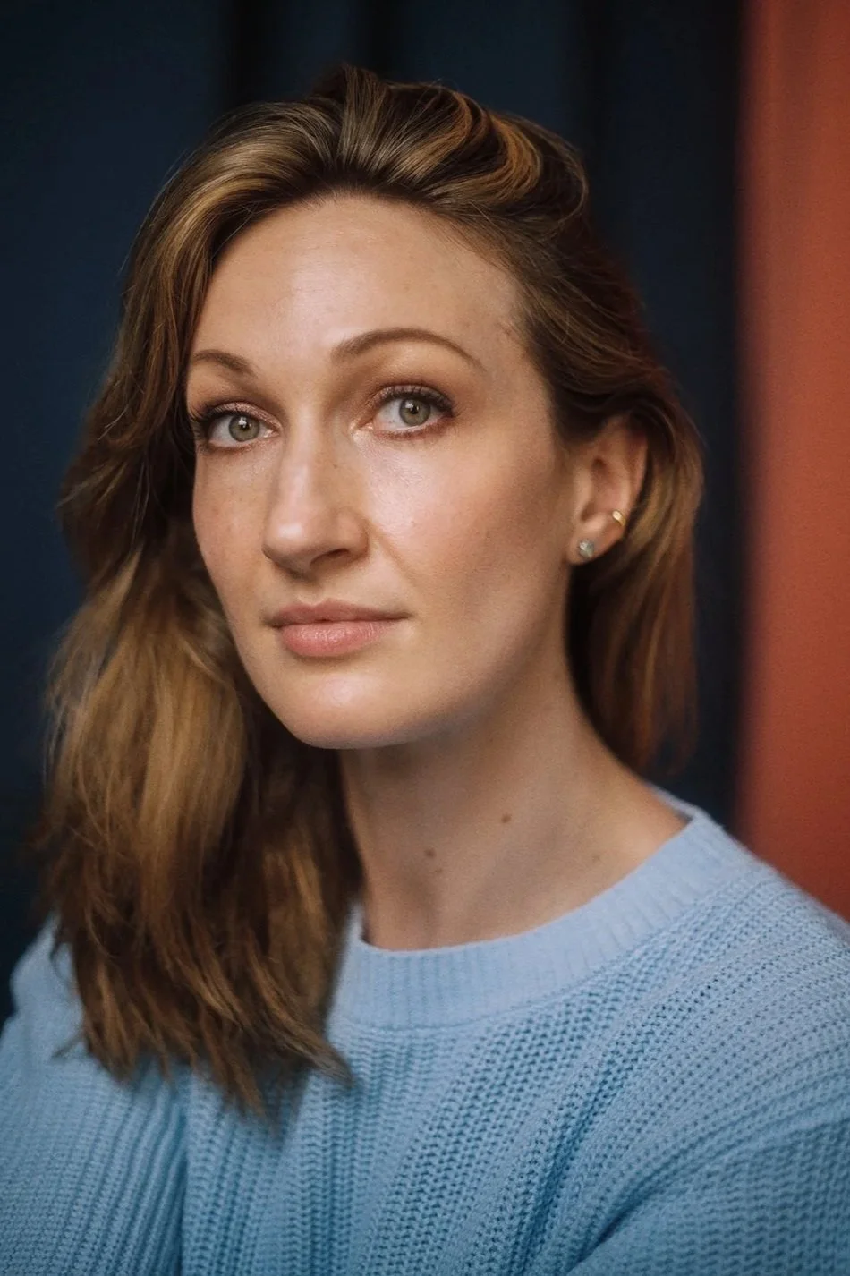 Close-up of a woman with light brown wavy hair, green eyes, and light skin, wearing a light blue sweater and small earrings, against a dark background with a reddish wall on the right.