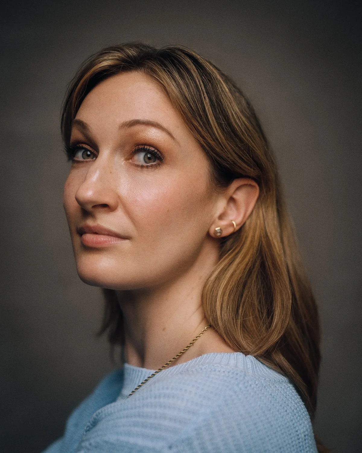 Close-up portrait of a woman with light brown hair, wearing a light blue top, small gold earrings, and a gold chain necklace, against a neutral dark background.