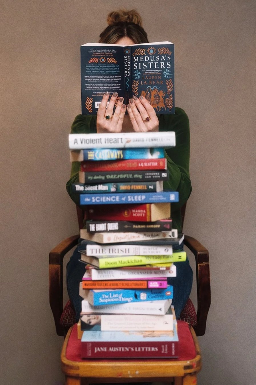 Person sitting on a wooden chair holding a book titled 'Medusa's Sisters' in front of their face, with a stack of various books covering their body and lap, against a plain background.