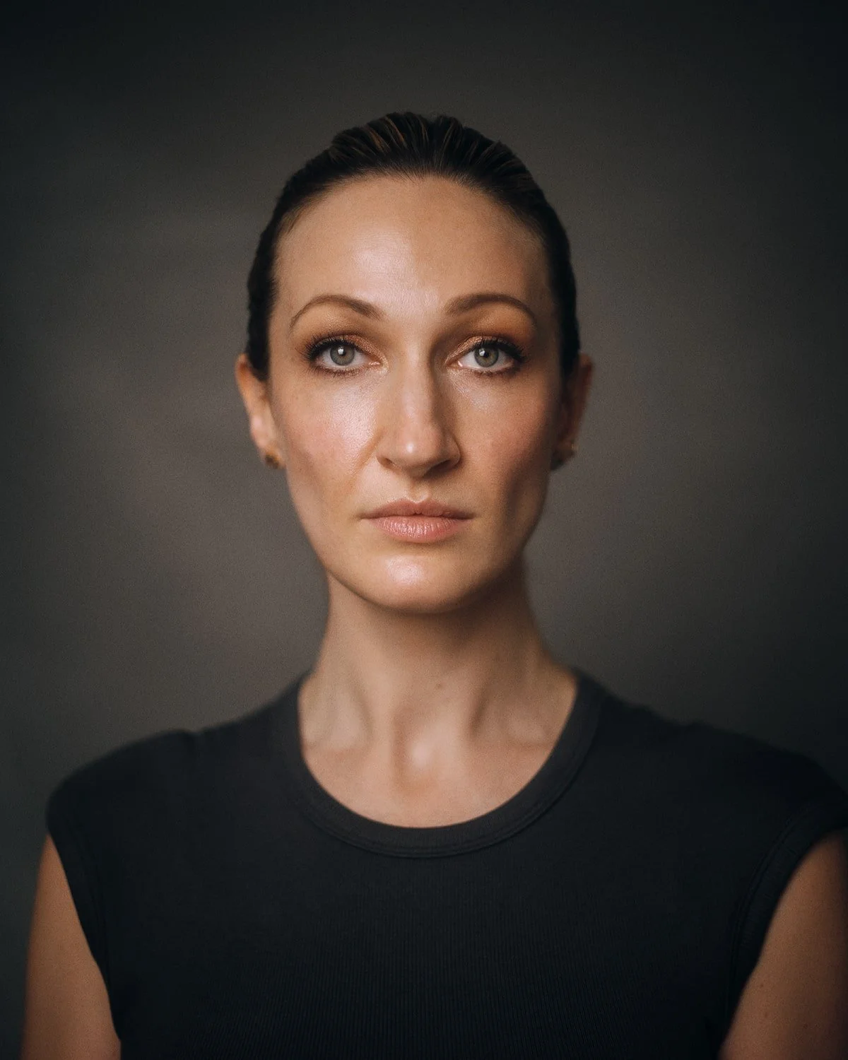 Portrait of a woman with slicked-back hair, wearing a black sleeveless top, against a dark background.