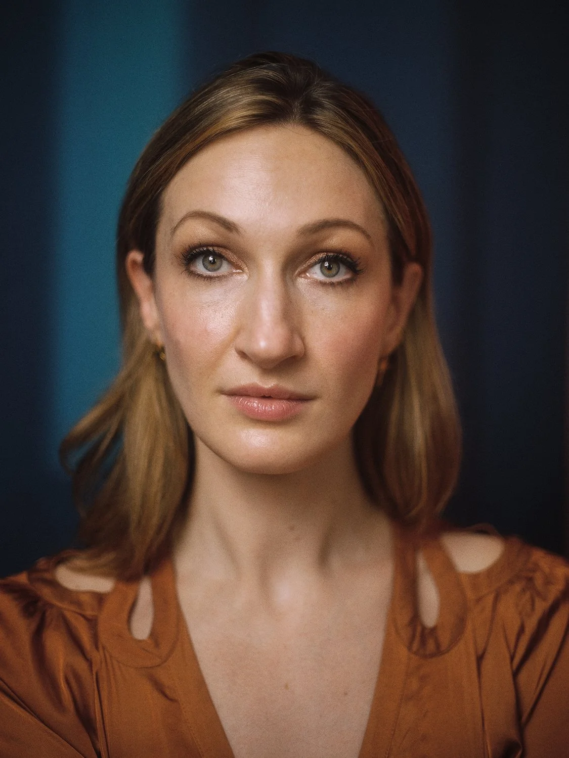 Close-up portrait of a woman with light brown hair, fair skin, and green eyes, wearing a brown blouse with cutout shoulders, against a dark background.