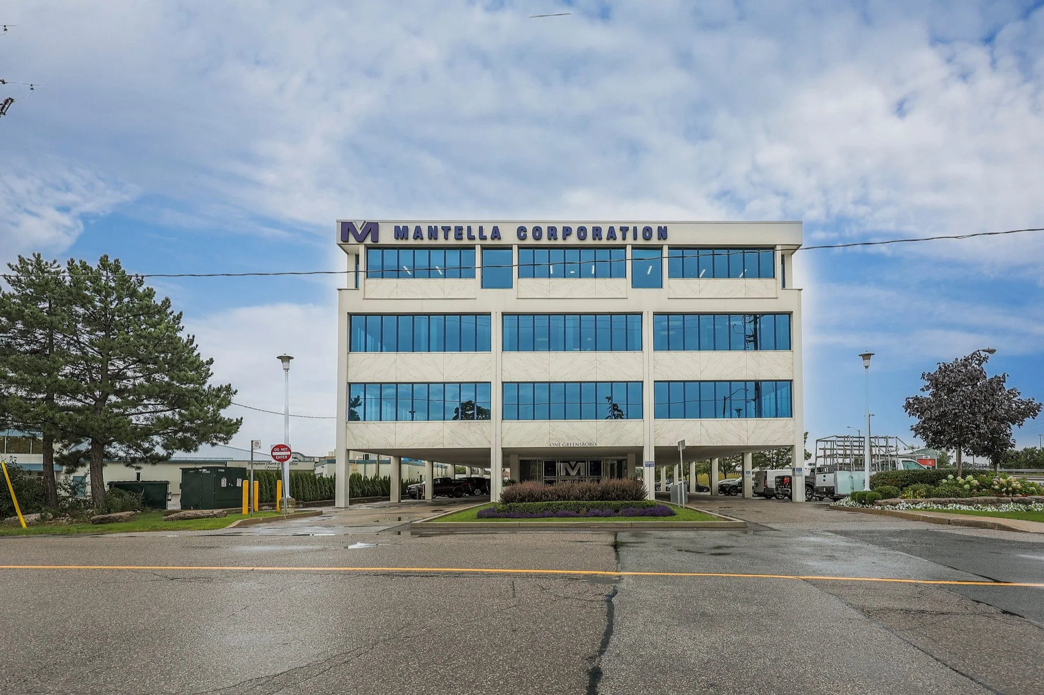 Front view of a modern office building with the sign 'Mantella Corporation' on top, surrounded by trees and parked vehicles under a partly cloudy sky.