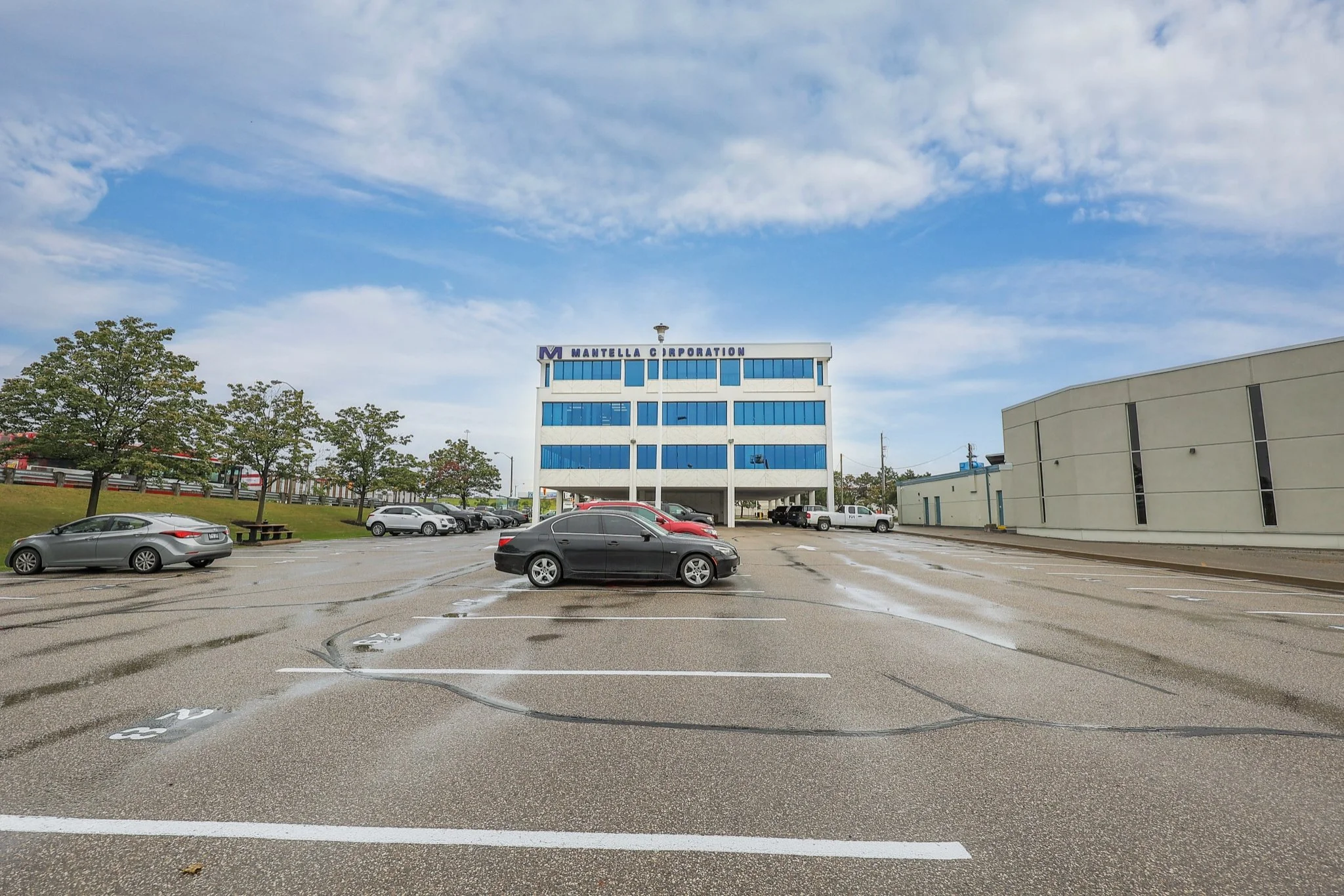 A parking lot with several parked cars and a building with blue windows and a sign that reads 'Mantella Corporation' under a partly cloudy sky.