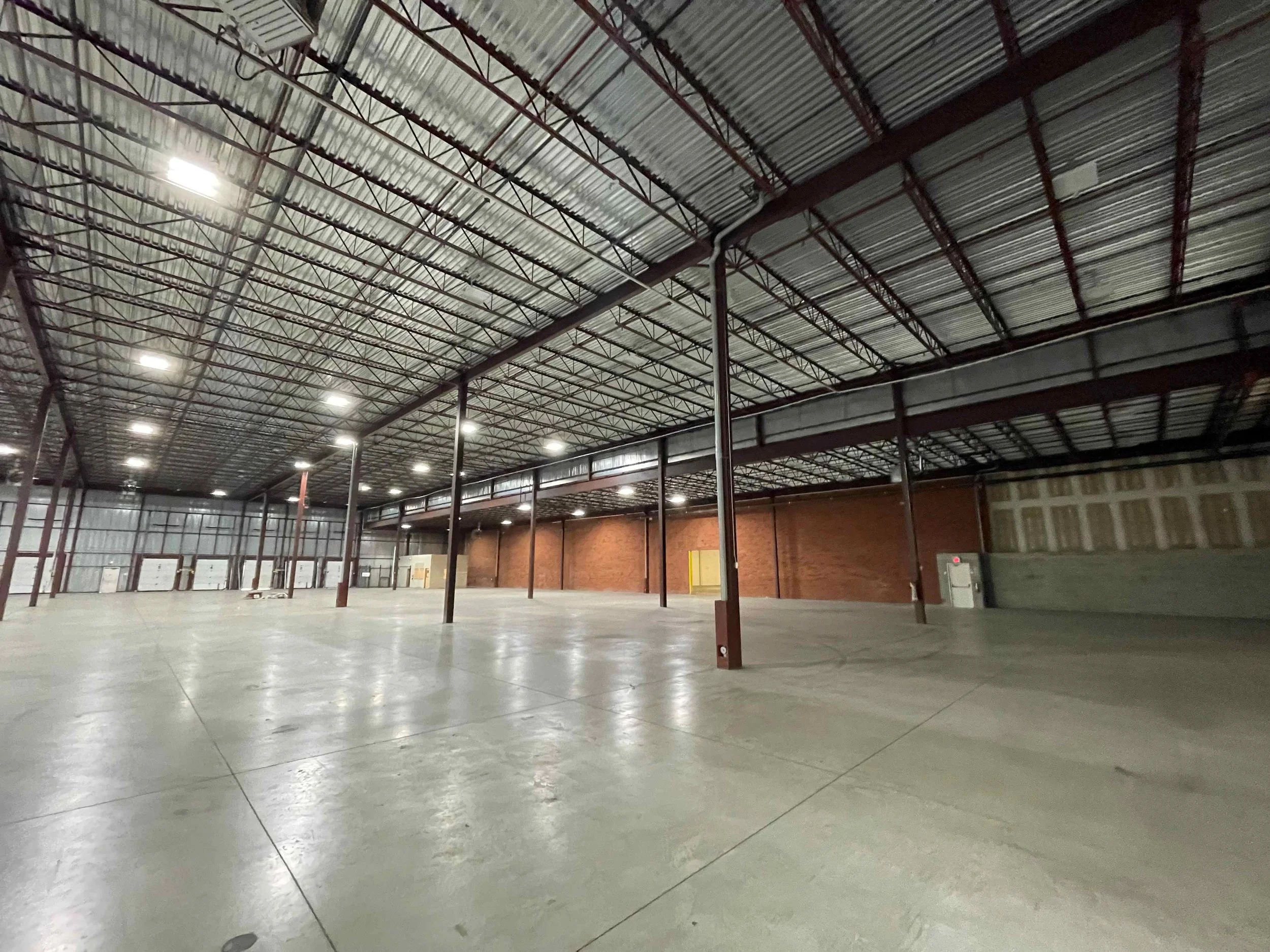 Interior of a large, empty industrial warehouse with a high metal ceiling, exposed beams, bright overhead lights, and a smooth concrete floor.