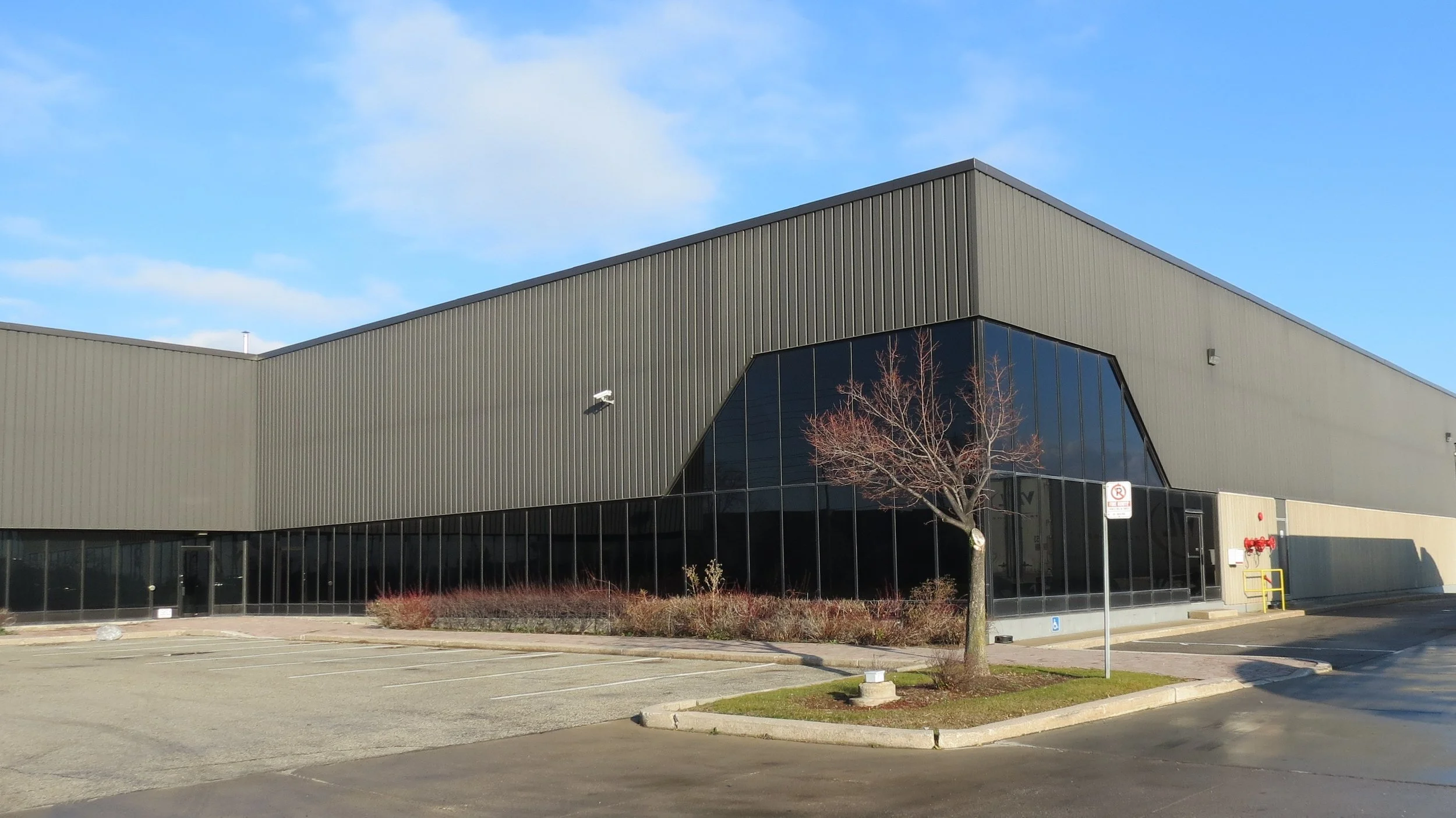 A large industrial-style building with grey metal siding and large glass windows, a few leafless trees in front, and a parking lot in the foreground under a clear blue sky.