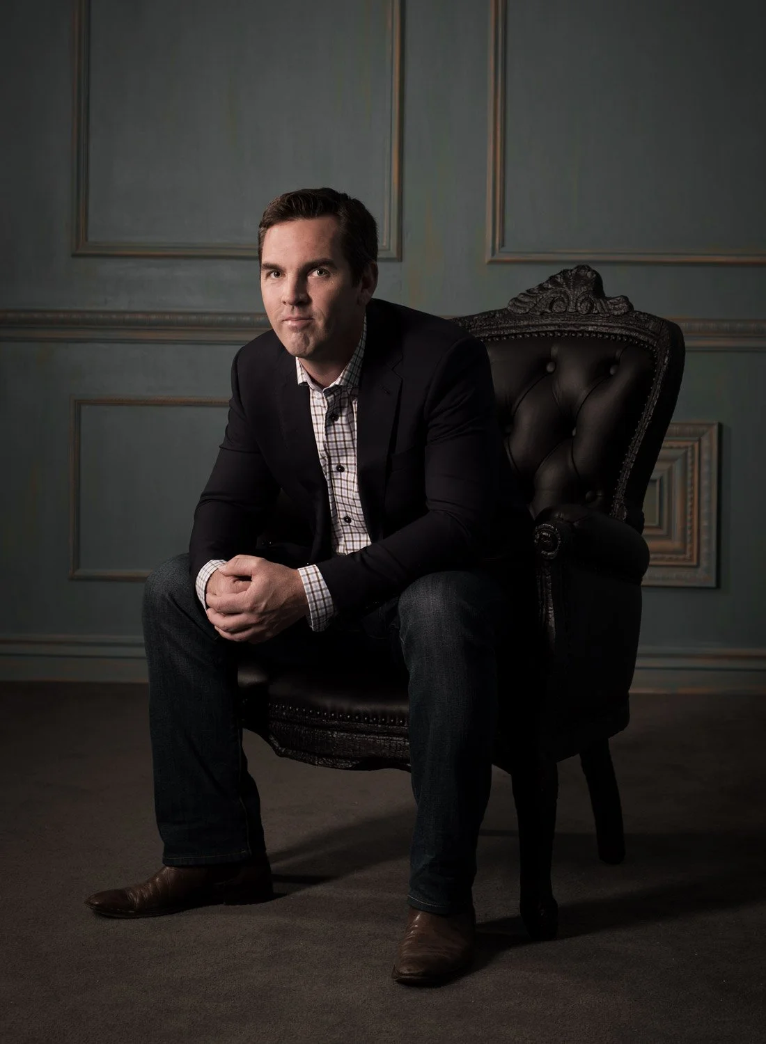 A man in a dark suit and checkered shirt sitting on a black upholstered armchair in a room with decorative wall panels.