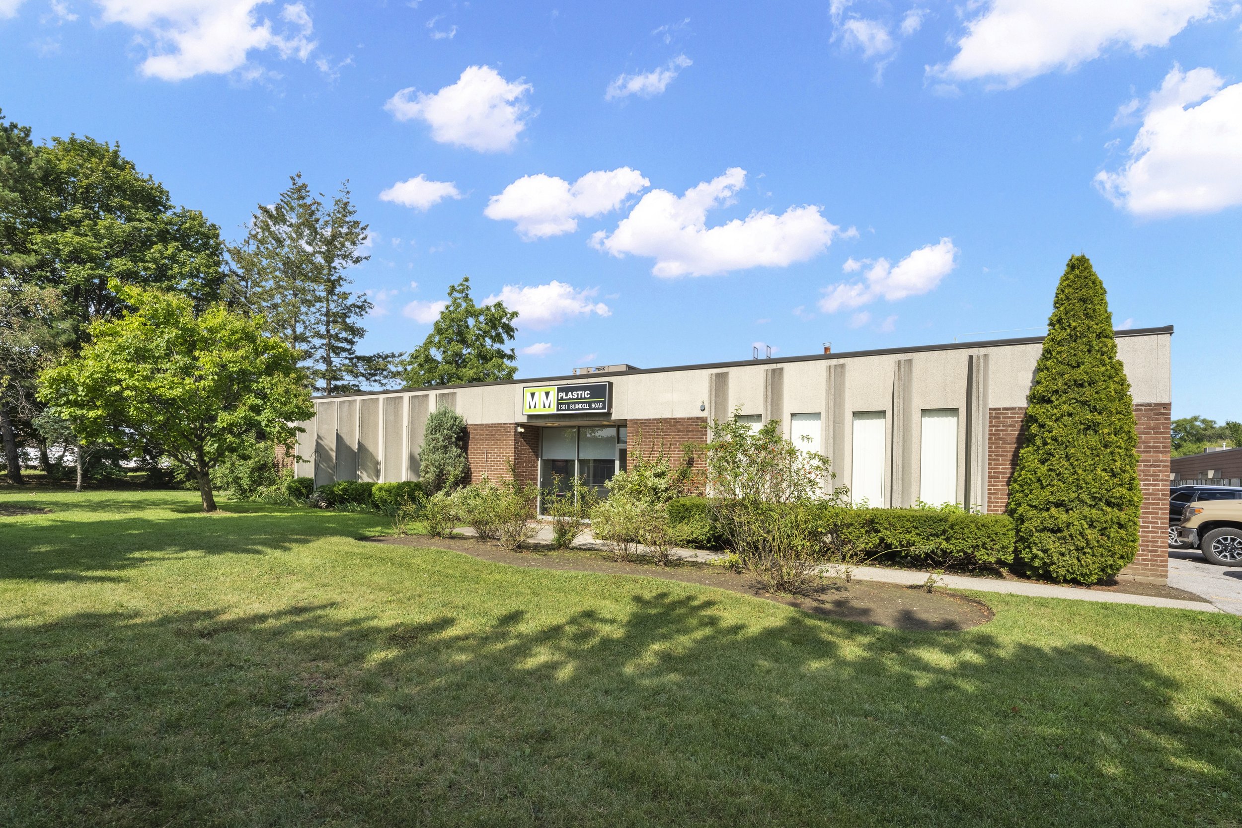A commercial building with a sign that reads 'MM Plastic,' surrounded by green trees and grass under a blue sky with white clouds.