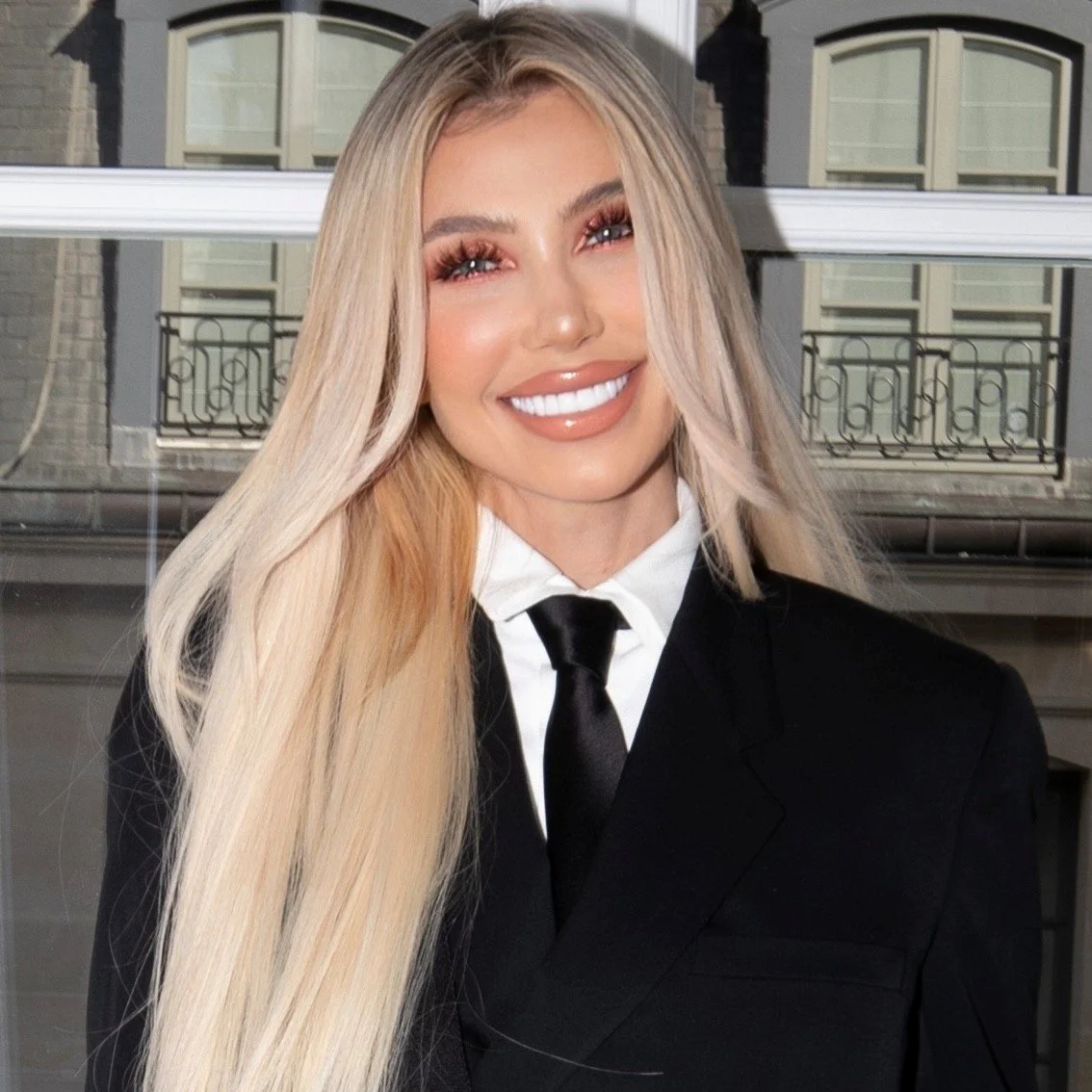 A woman with long blonde hair, wearing a black suit with a white shirt and black tie, smiling with visible teeth, in front of a building with windows and balconies.
