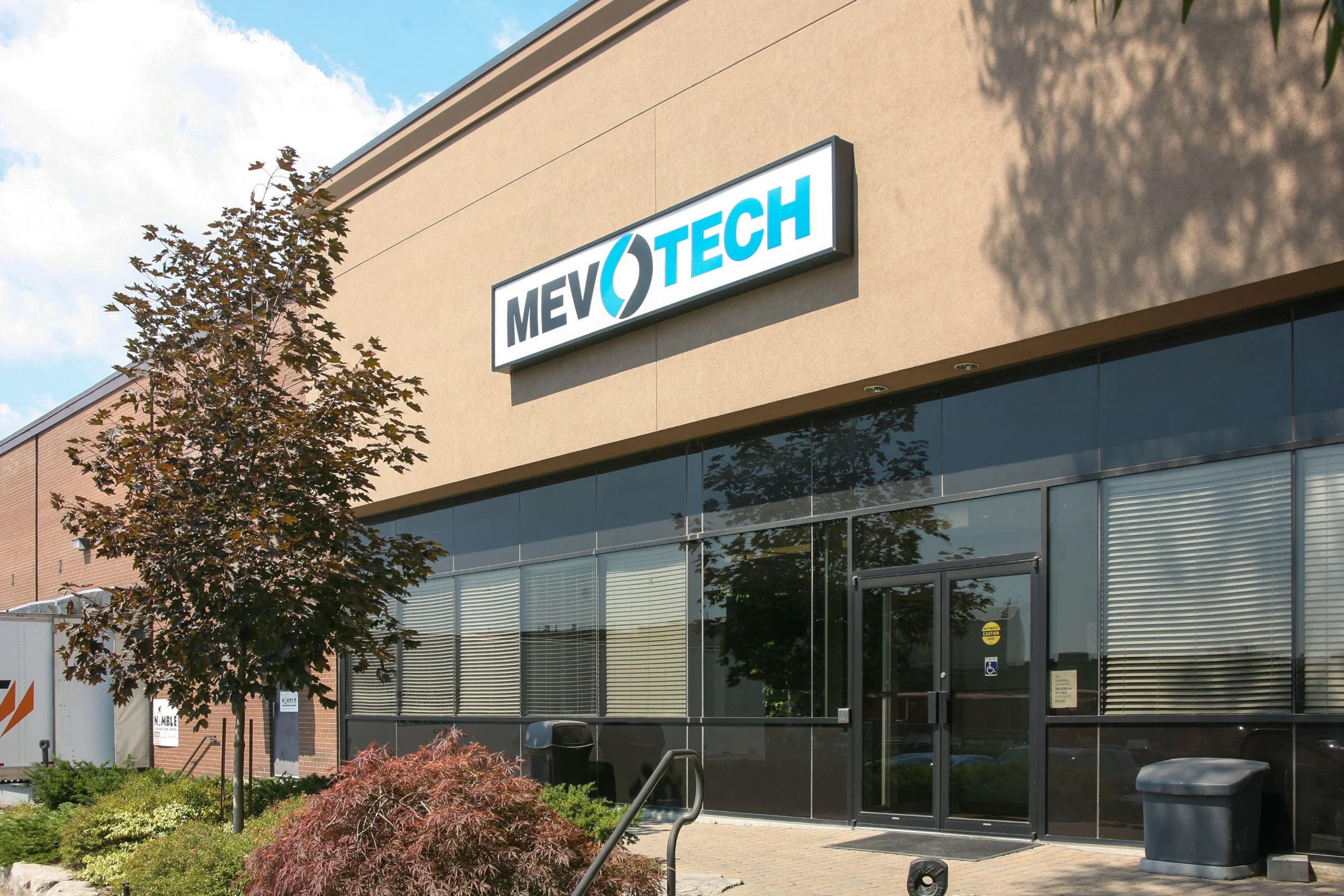 Exterior of a commercial building with a sign that reads "MEVO TECH." The building has a beige upper wall and a lower section with large glass windows. There are trees and shrubs in the foreground, and the sky is partly cloudy.