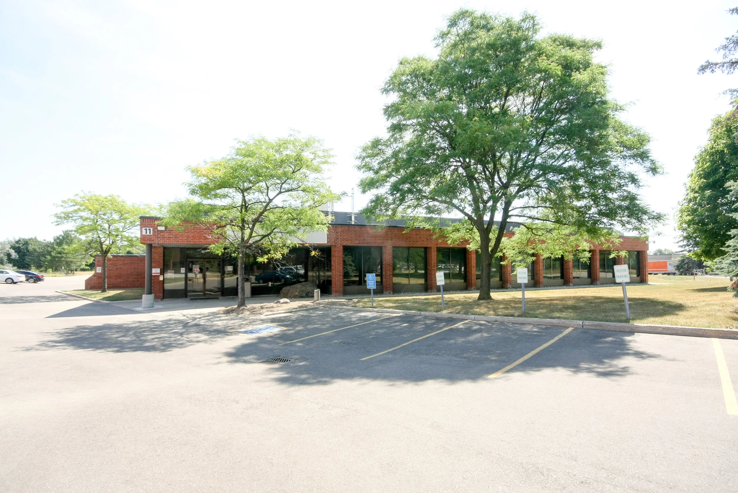 A parking lot with several marked parking spaces, some with cars, and a brick building with large glass windows behind two trees. There are signs indicating visitor parking and a handicapped parking space, along with some large rocks near the trees.