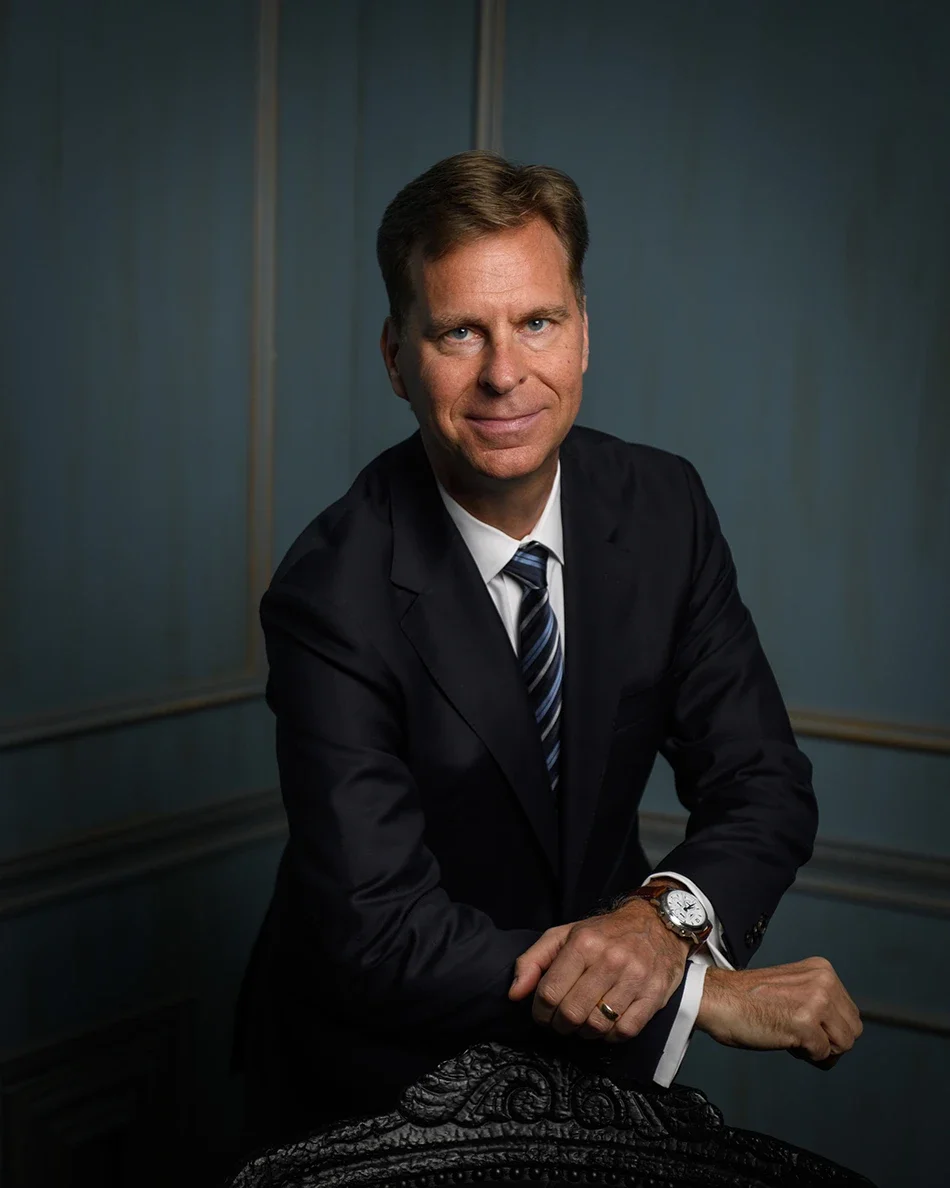 A man with light brown hair in a black suit and white shirt sitting on a black leather chair in a room with blue dark grey/blue walls..