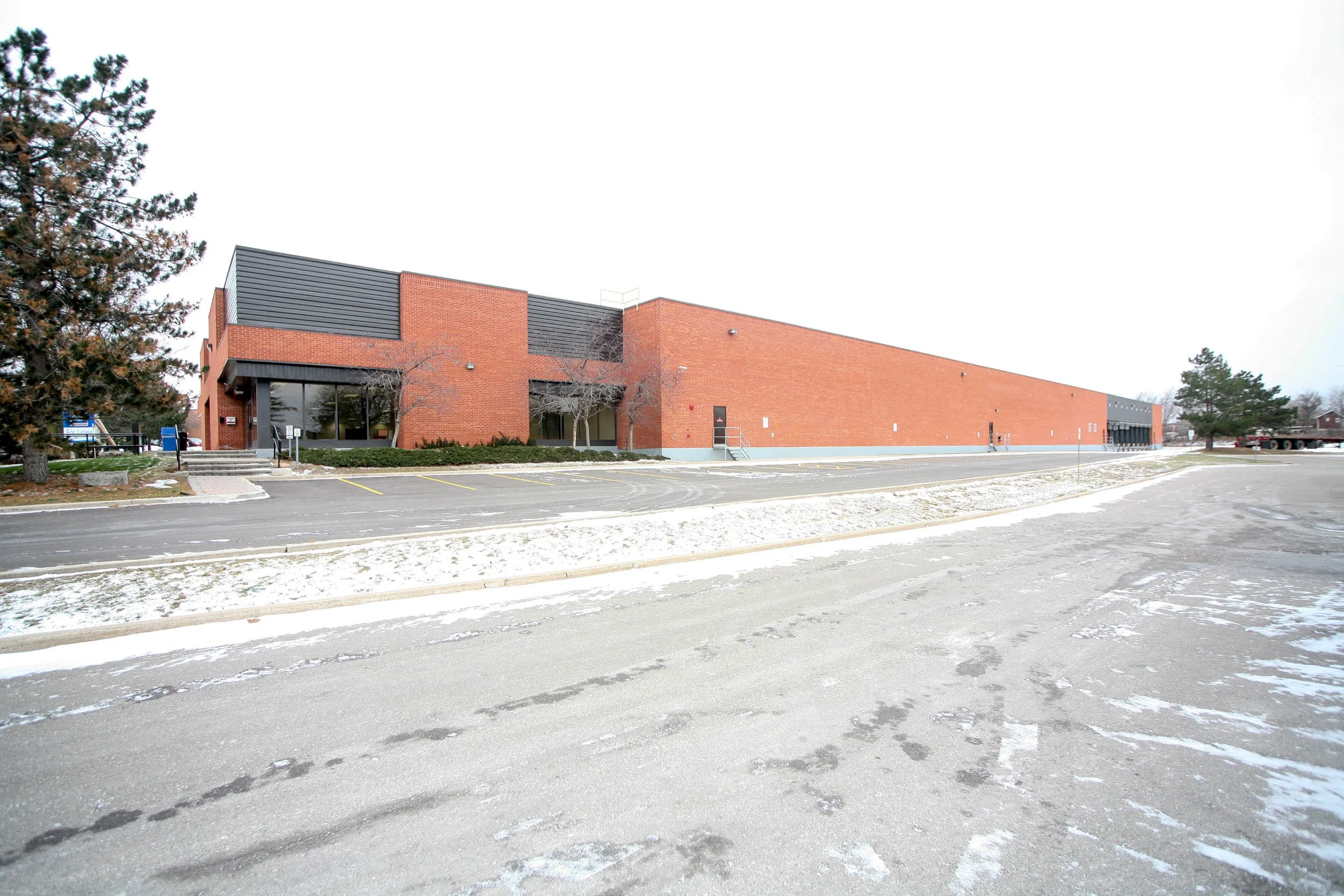 A modern brick building with large windows and an overcast sky, surrounded by a parking lot with some snow and bare trees.