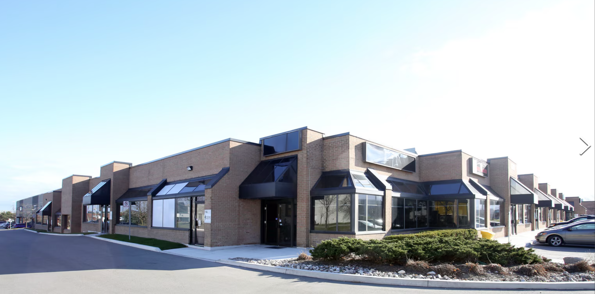 A modern commercial building with large glass windows and black awnings, situated along a paved road with parking spaces and some cars, under a clear blue sky.