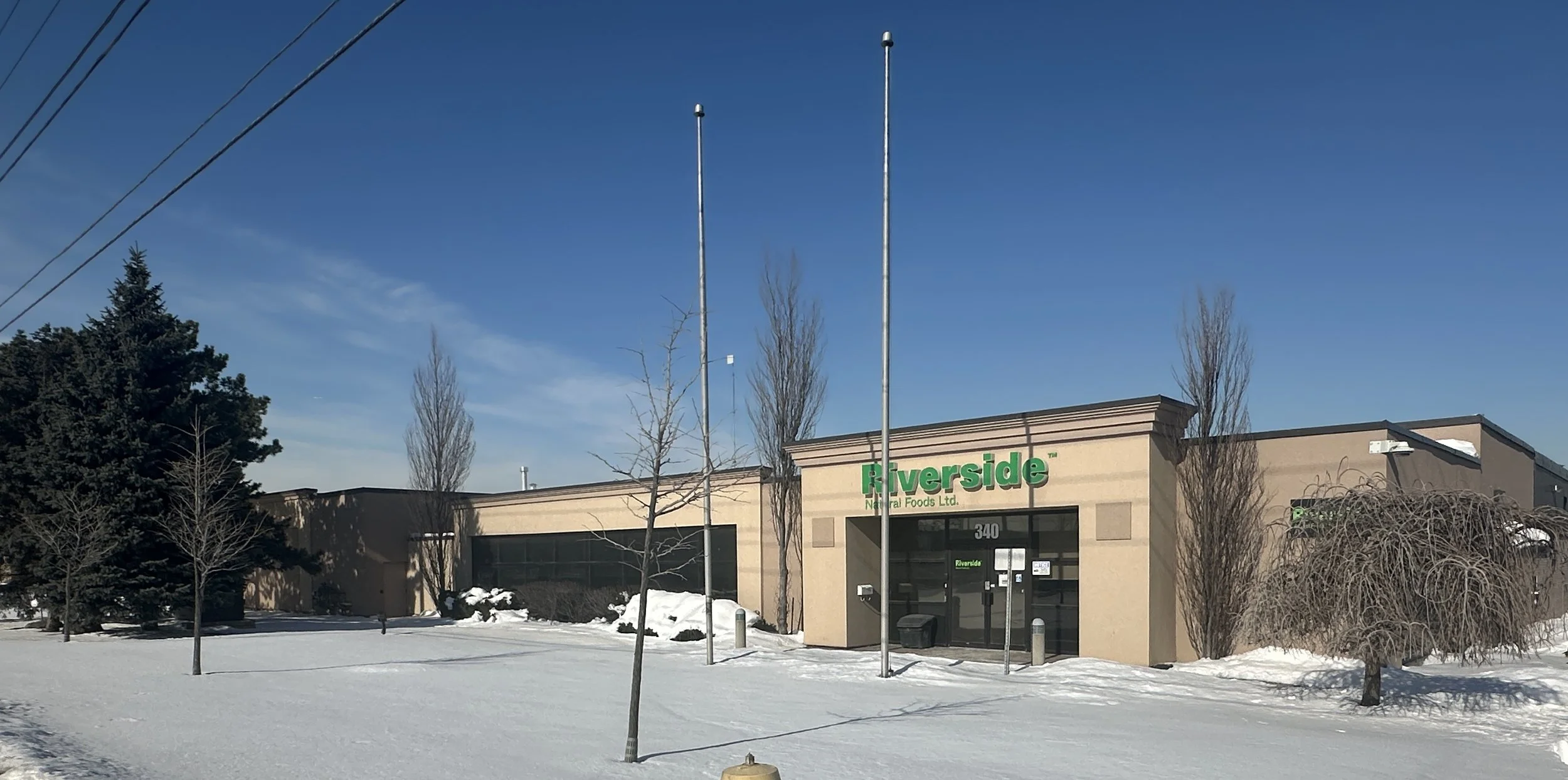 A single-story beige commercial building with a green Riverside sign, surrounded by leafless trees and snow-covered ground, under a clear blue sky.
