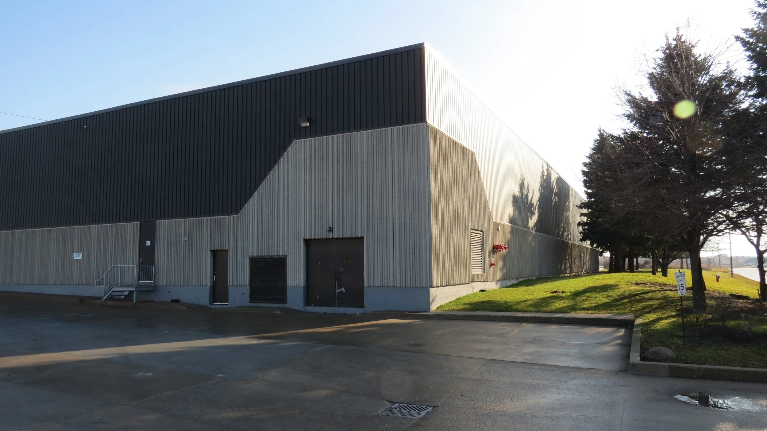 A large industrial building with a gray exterior, black upper section, and multiple doors. There are trees and a grassy area beside the building, with a speed limit sign indicating 15 mph.