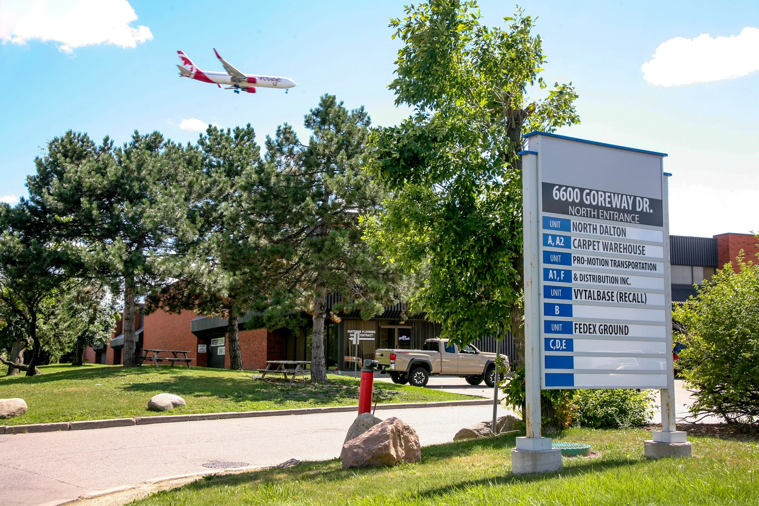 A parking lot with a sign listing different unit locations, trees, a parked pickup truck, and an airplane flying in the clear blue sky.
