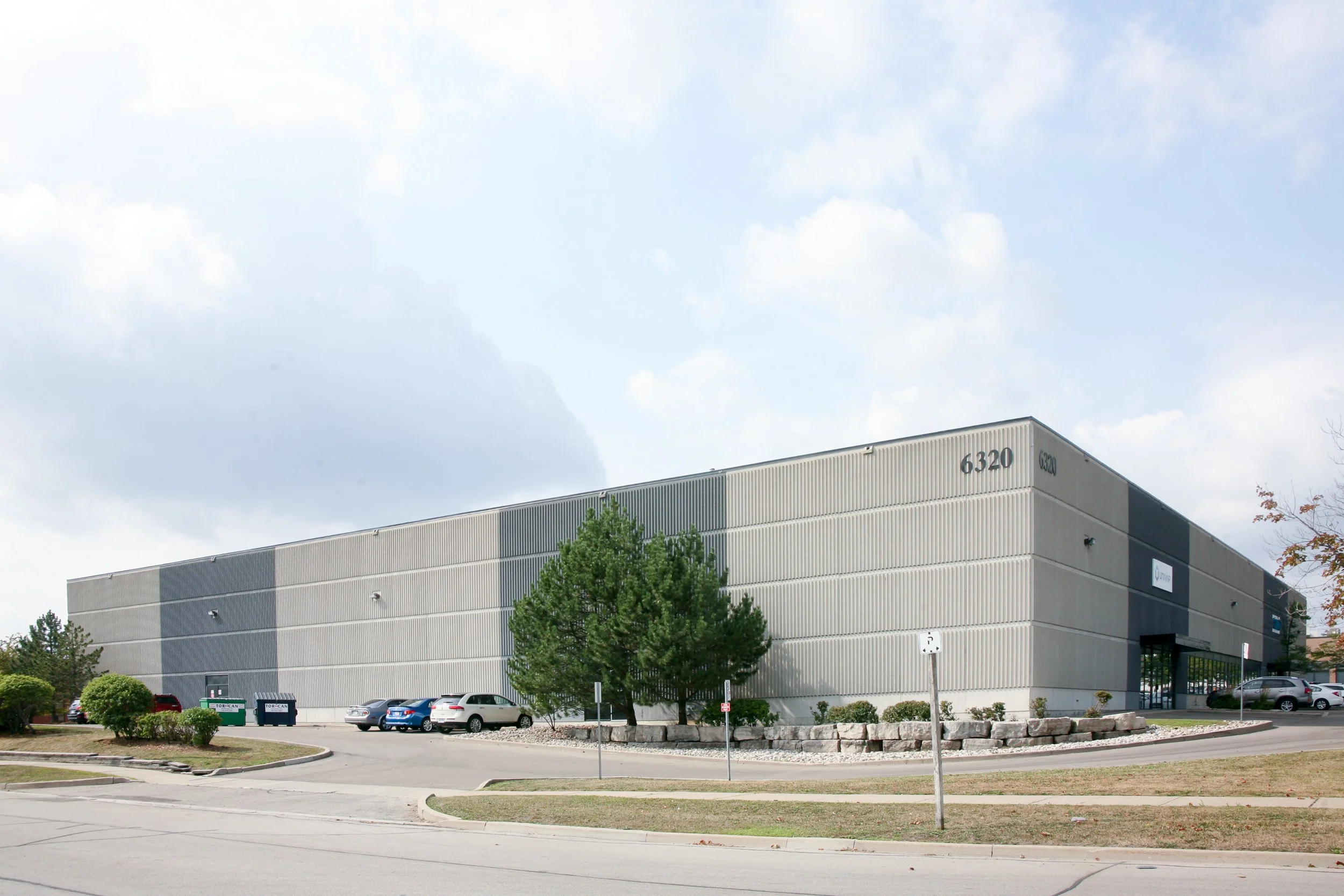 A large, modern commercial building with a gray exterior, several parking spaces filled with cars, and trees in front. The sky above is partly cloudy.