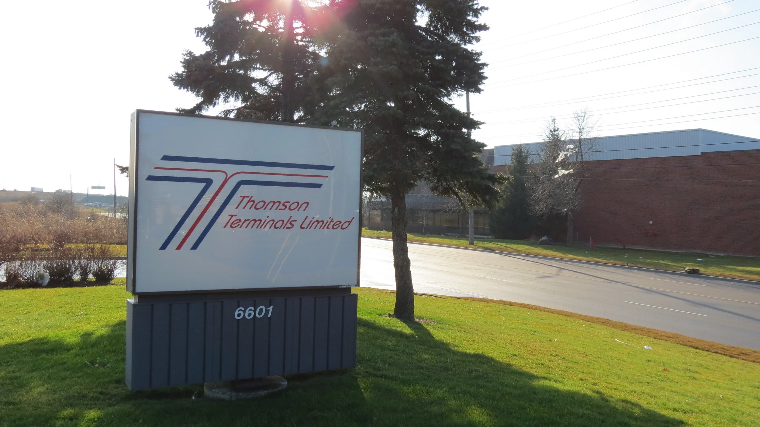 Sign for Thomson Terminals Limited with the address 6601, near a tree, paved road, and industrial buildings in the background.