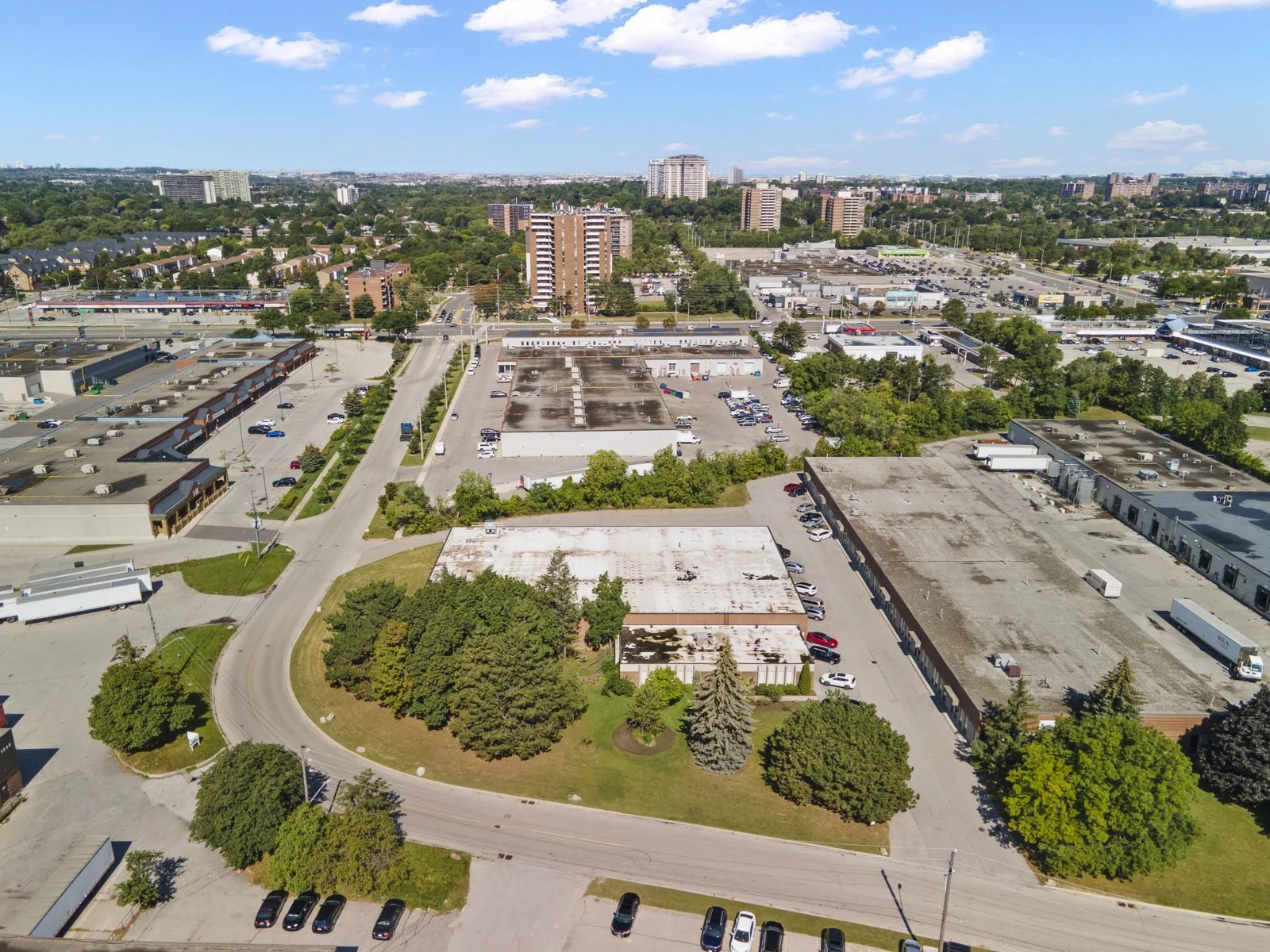 Aerial view of a cityscape with commercial buildings, parking lots, high-rise apartment towers, and green trees under a clear blue sky.