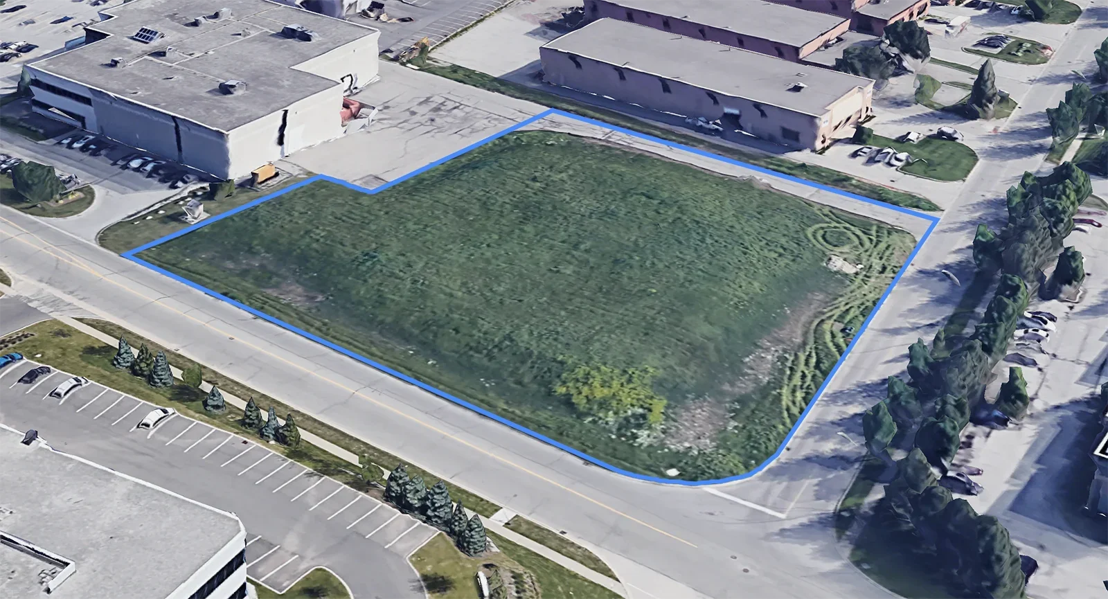 A vacant grassy lot outlined in blue, adjacent to parking lots and buildings, with trees along the street.