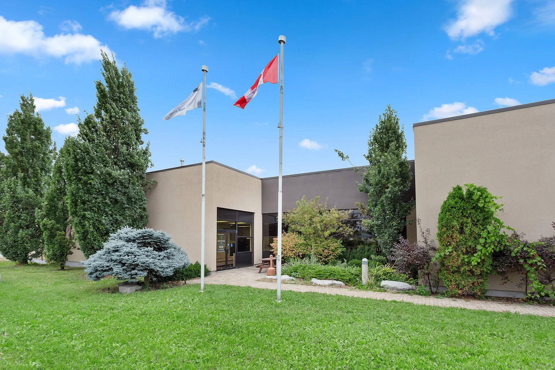 The exterior of a modern building with a landscaped garden, three flagpoles with flags, and a sunny blue sky with a few clouds.