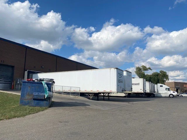 A row of white semi-trailers parked next to a warehouse on a paved lot with a blue sky and clouds overhead.