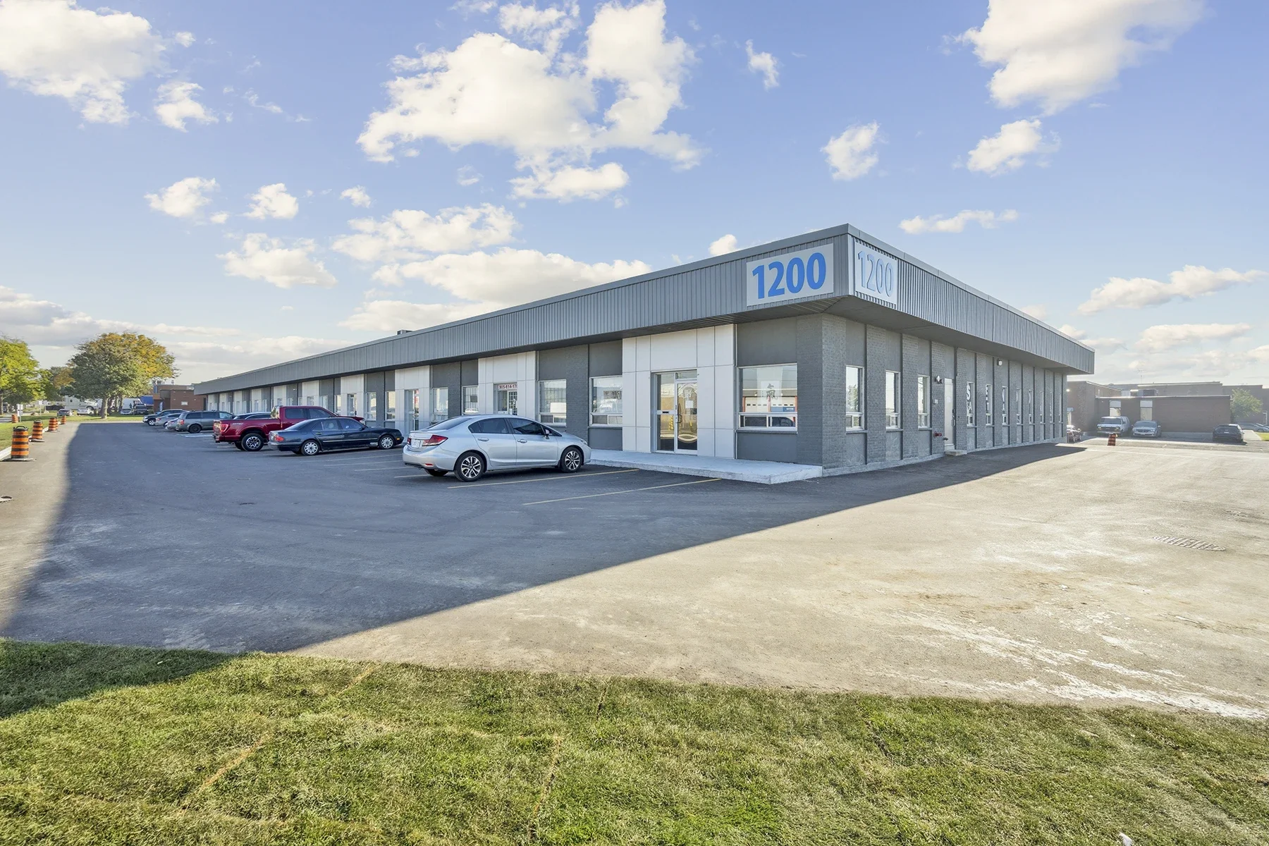 Commercial building with a large blue '1200' sign on the corner, parking lot with several cars, some trees, and a partly cloudy sky.
