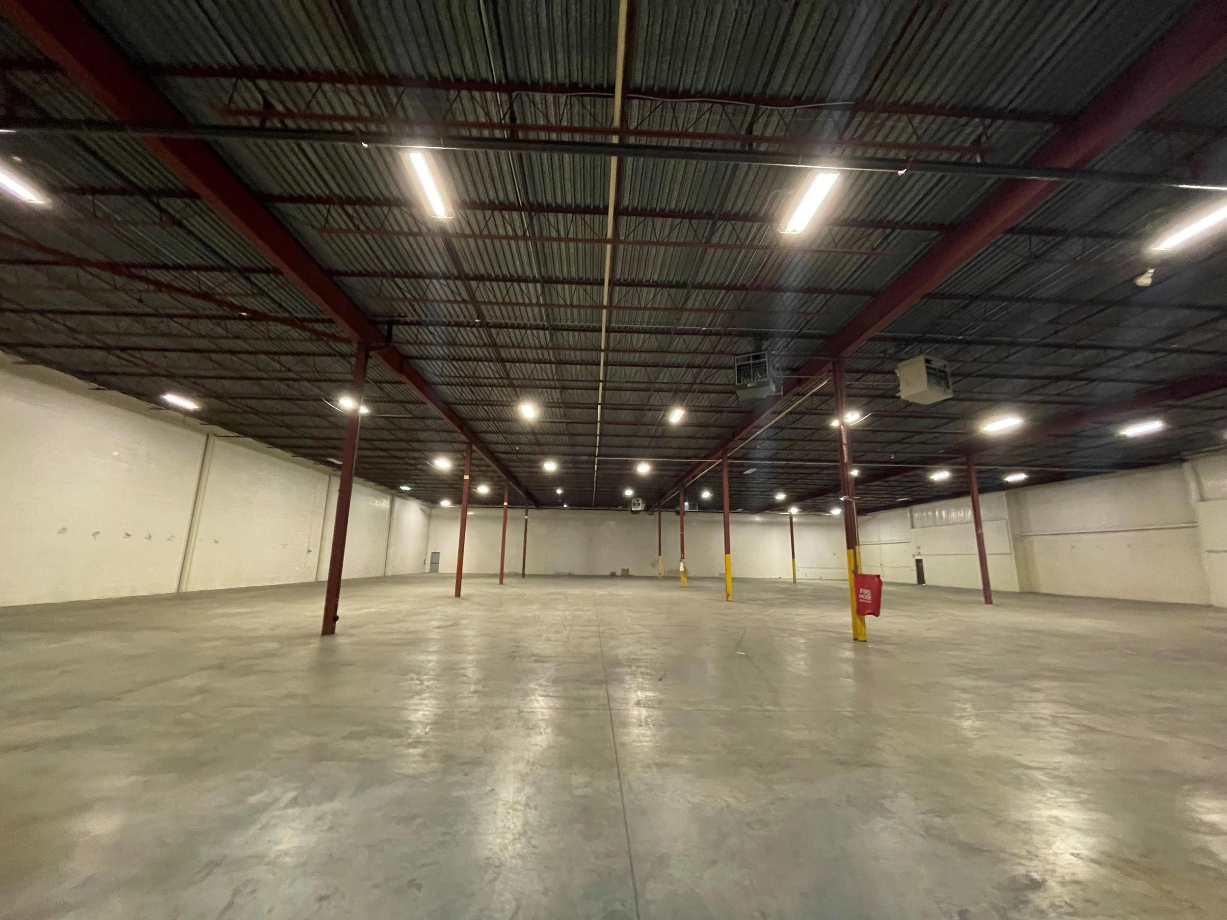 Empty warehouse with high metal ceiling, exposed red beams, and bright overhead lighting.