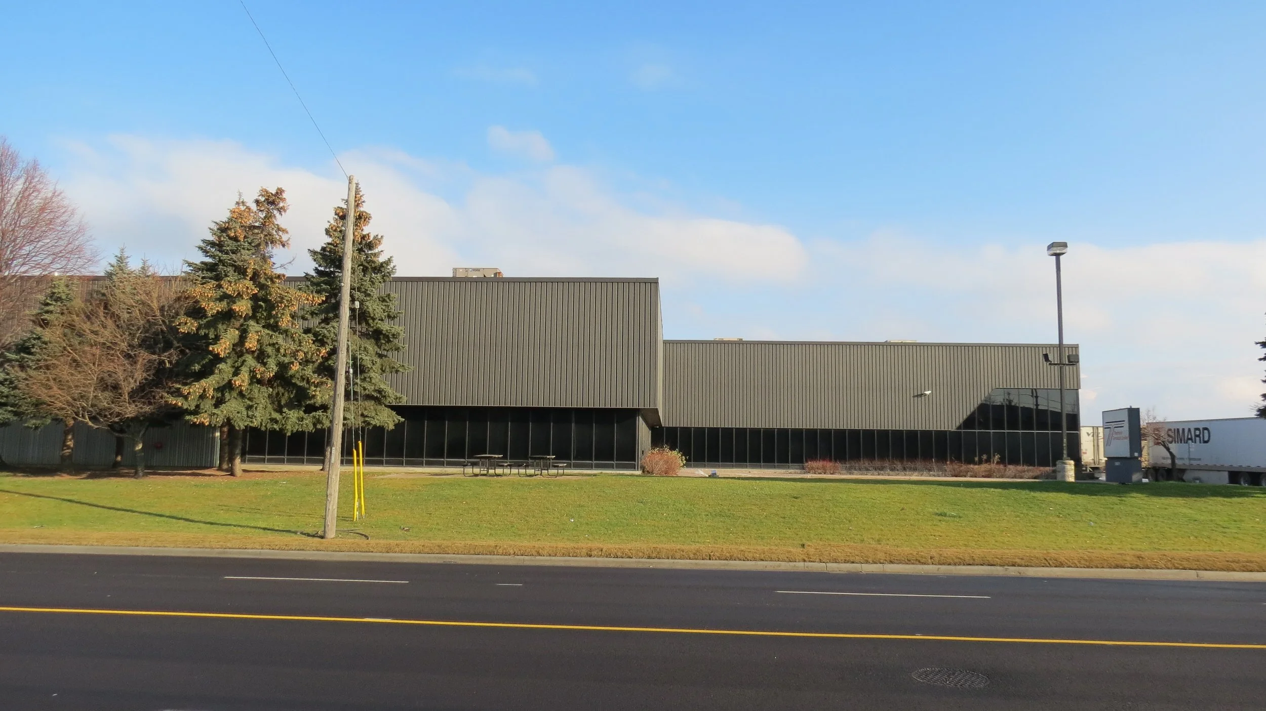 A commercial building with a gray metal exterior, steel-trimmed windows, and a side parking area with a large truck, situated along a street with a grassy area and trees, under a clear blue sky.