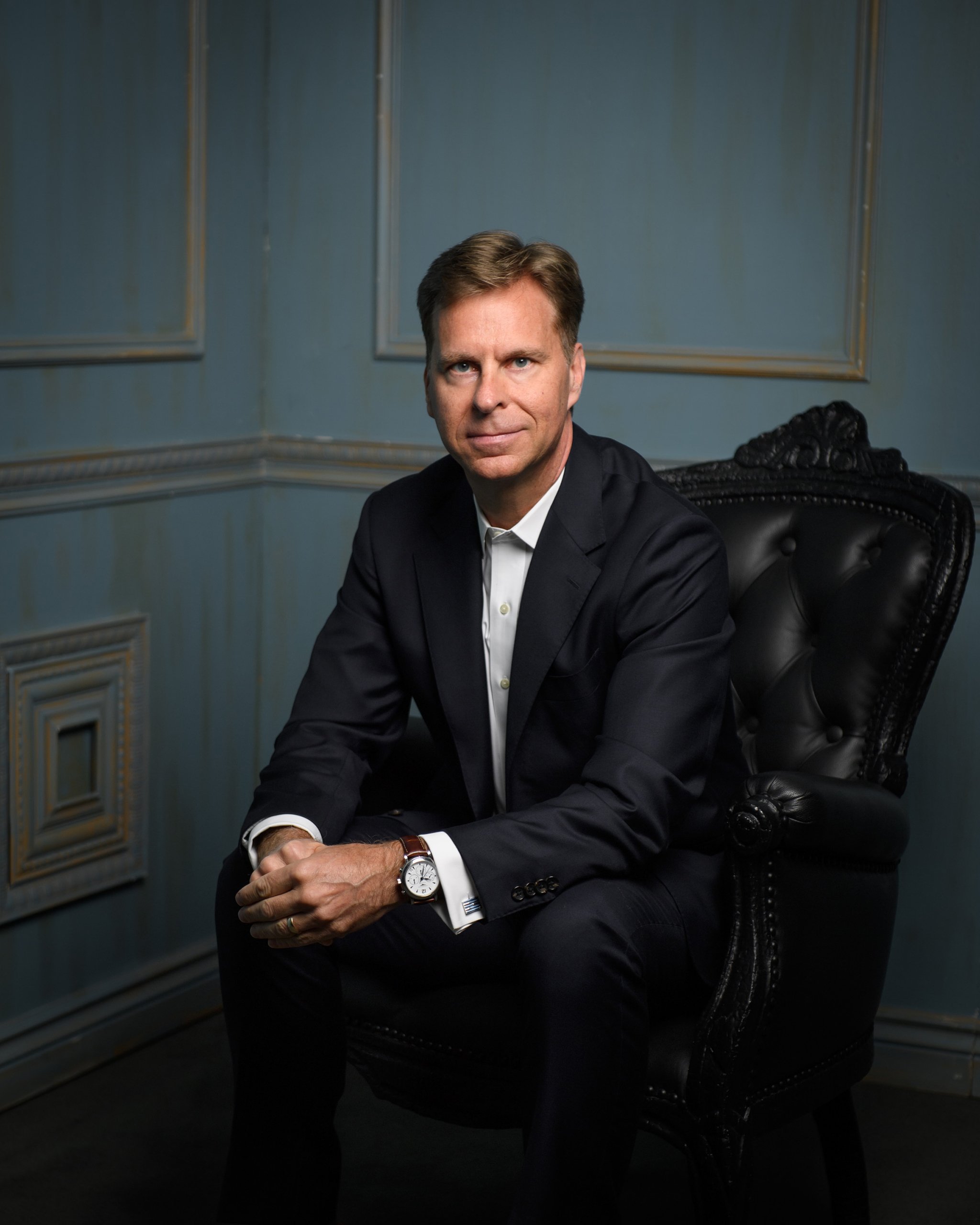 A man with light brown hair in a black suit and white shirt sitting on a black leather chair in a room with blue decor.