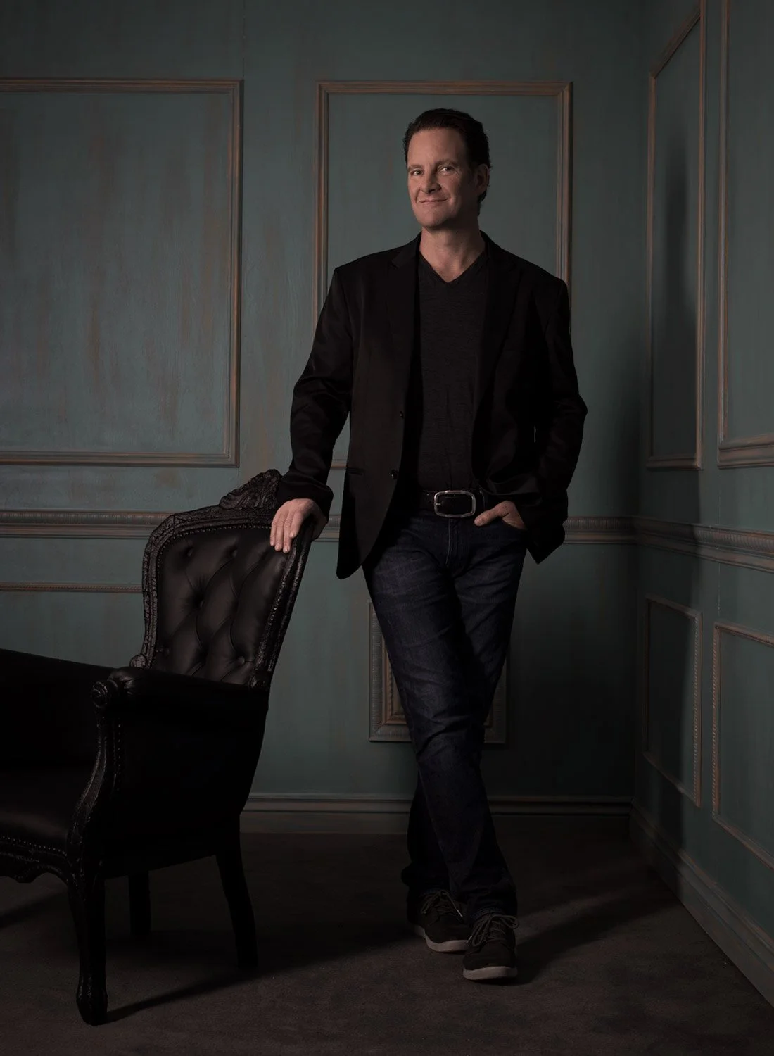 A man in a black blazer and jeans standing in a room with blue paneled walls, leaning on a black leather chair.