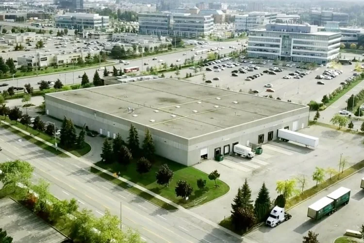 A large industrial building with loading docks, surrounded by parking lots, trees, and nearby office buildings.
