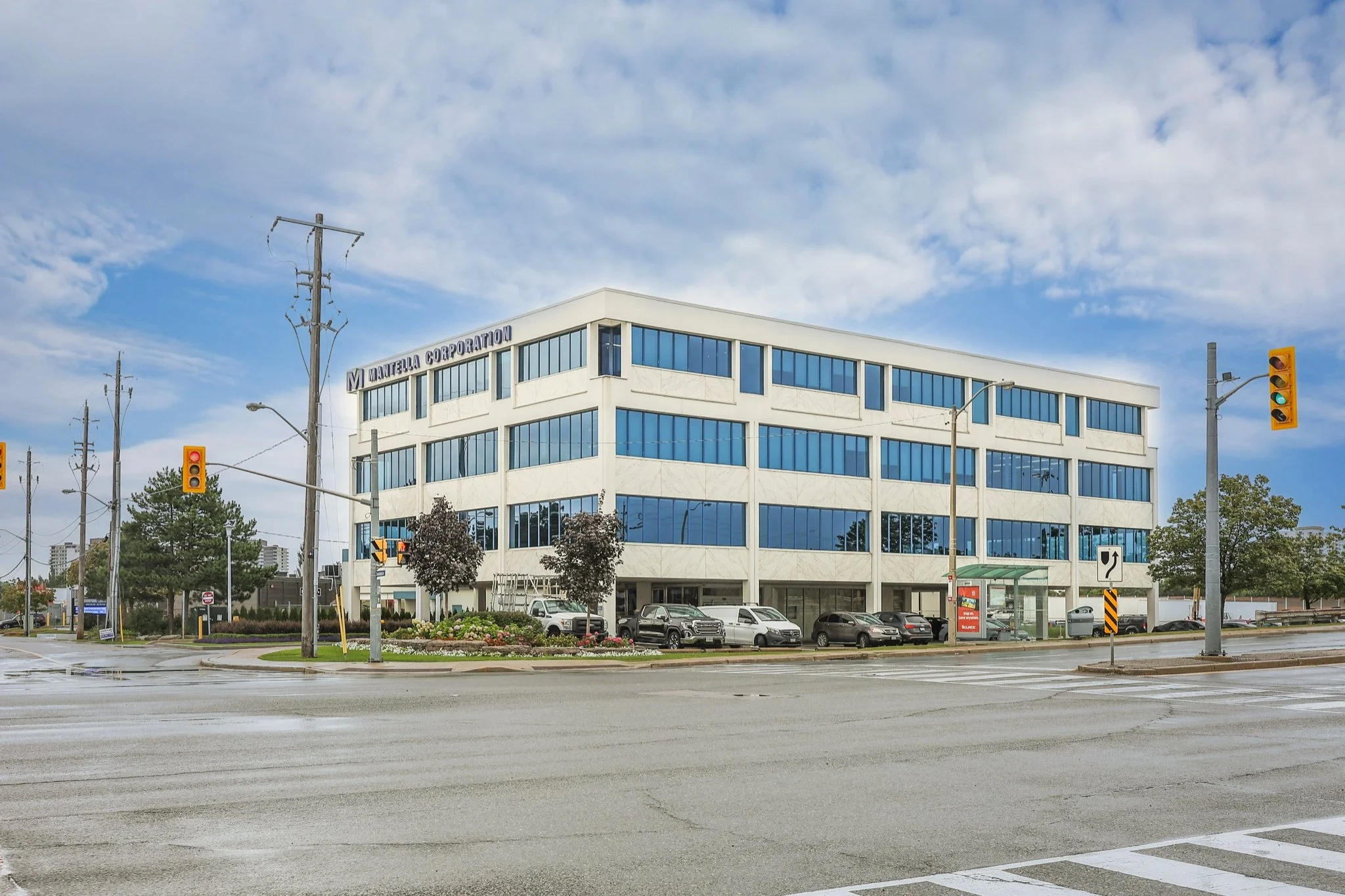 A modern white office building with many large blue-tinted windows, with a sign that reads 'Matrella Corporation' on top. The street in front has traffic lights, cars parked along the curb, and some trees. The sky is partly cloudy.
