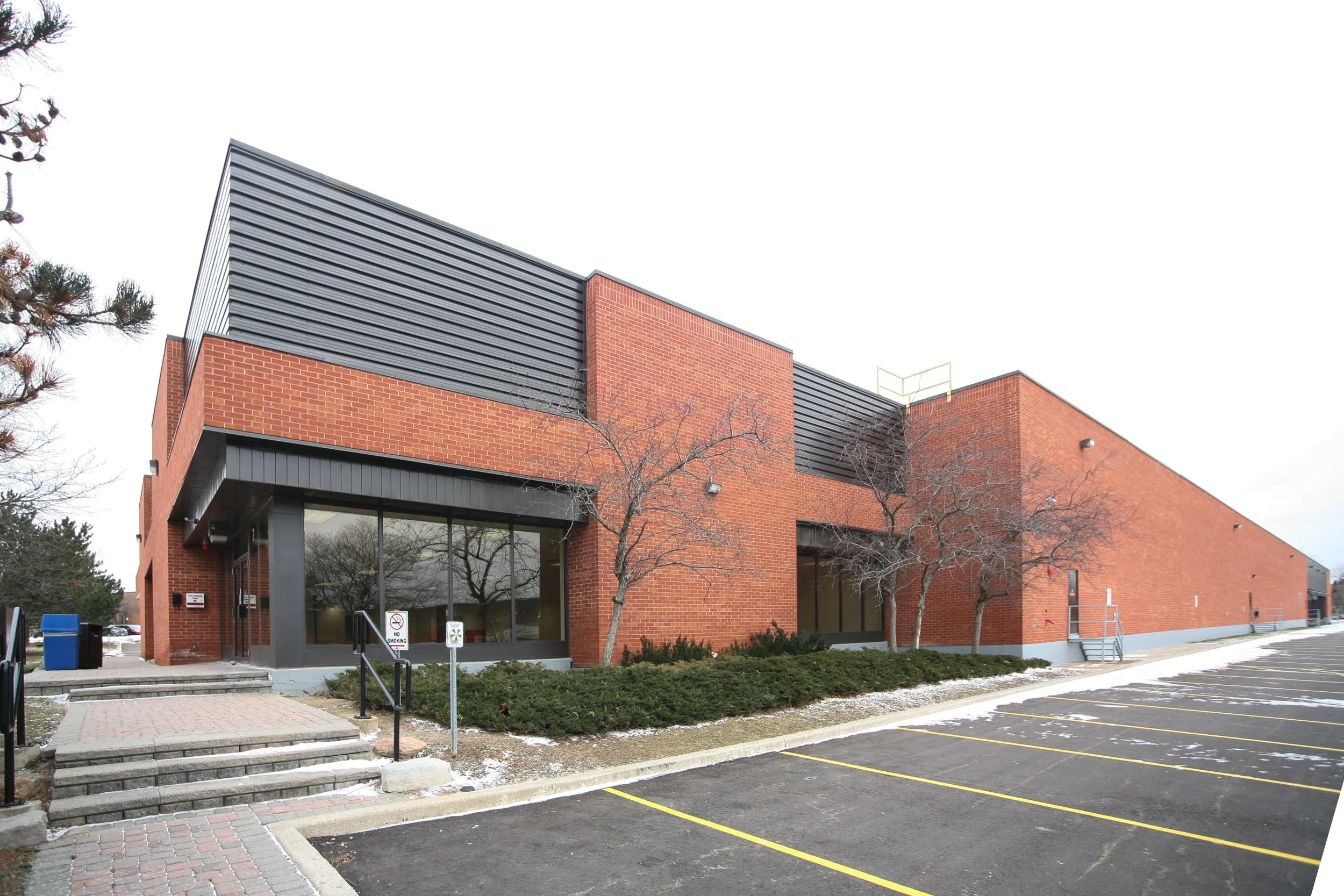 A modern brick building with black metal accents, large glass windows, and a metal roof, with a parking lot in the foreground and leafless trees outside.