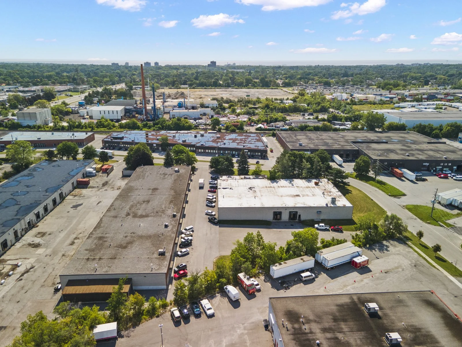 Aerial view of an industrial and commercial area with multiple buildings, parking lots, and trucks, surrounded by green trees and distant cityscape in the background.