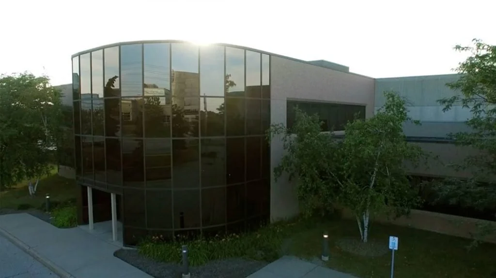 Modern building with curved glass facade and surrounding trees, sunlight shining on the top of the building.