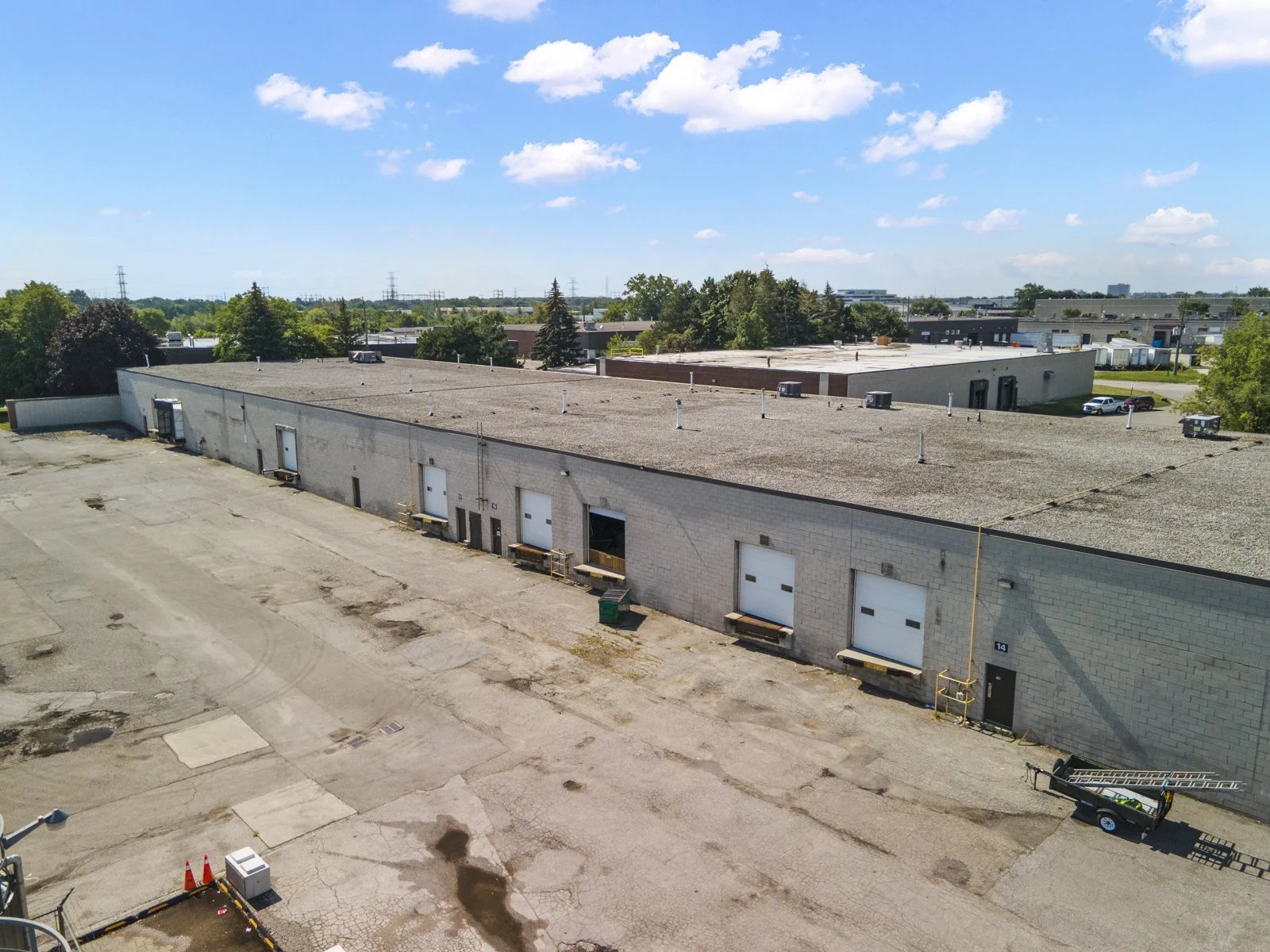 An aerial view of a large industrial building with a flat roof and multiple loading dock doors, surrounded by an empty parking lot and a few parked vehicles under a partly cloudy sky.