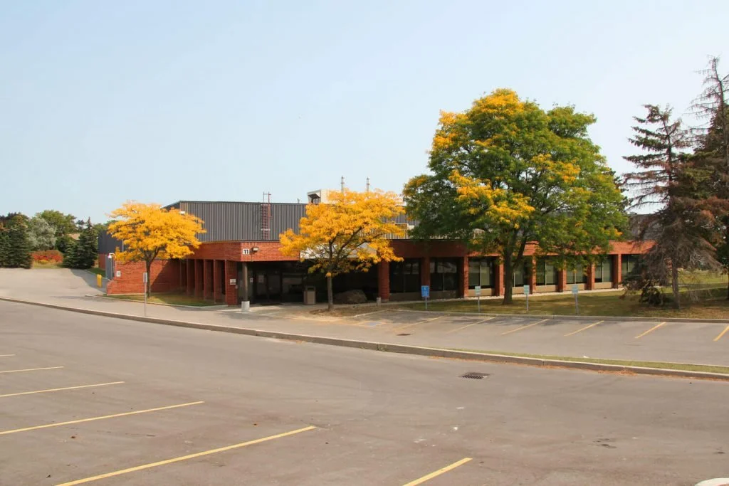 A brick building with a black roof surrounded by trees with yellow and green leaves, situated next to an empty parking lot on a cloudy day.