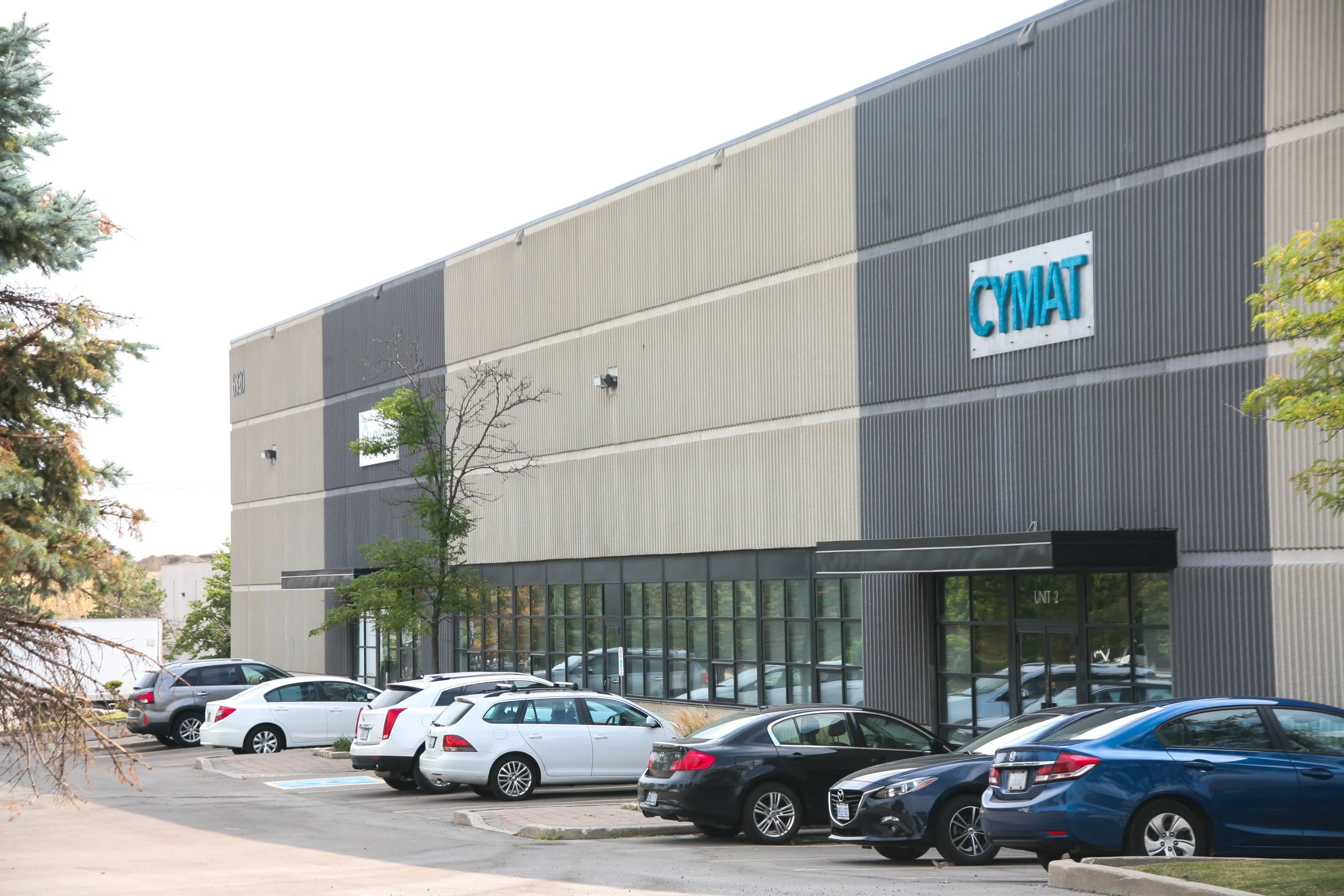 Parking lot in front of a commercial building with a sign labeled CYMAT, with several cars parked and trees nearby.