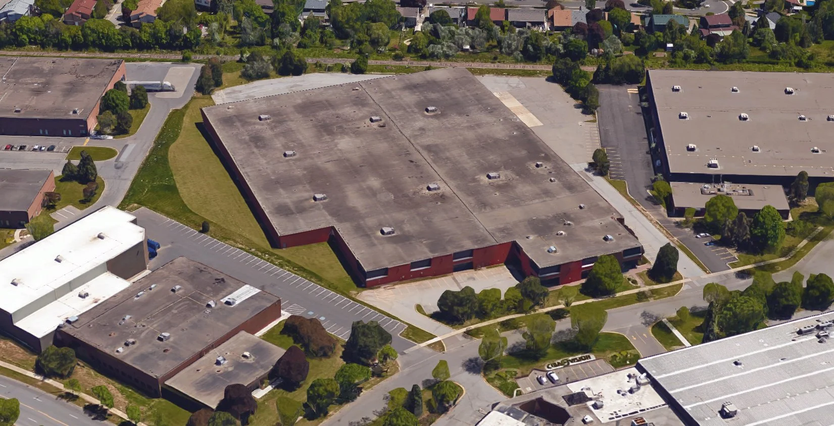 Aerial view of a large, flat-roofed industrial or commercial building surrounded by parking lots and greenery, located in a business or commercial area with residential houses in the background.
