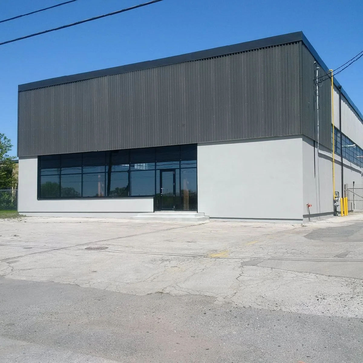 A modern two-story commercial building with a black metal upper section and white lower section, featuring large glass windows at the front, situated on a cracked parking lot under a clear blue sky.