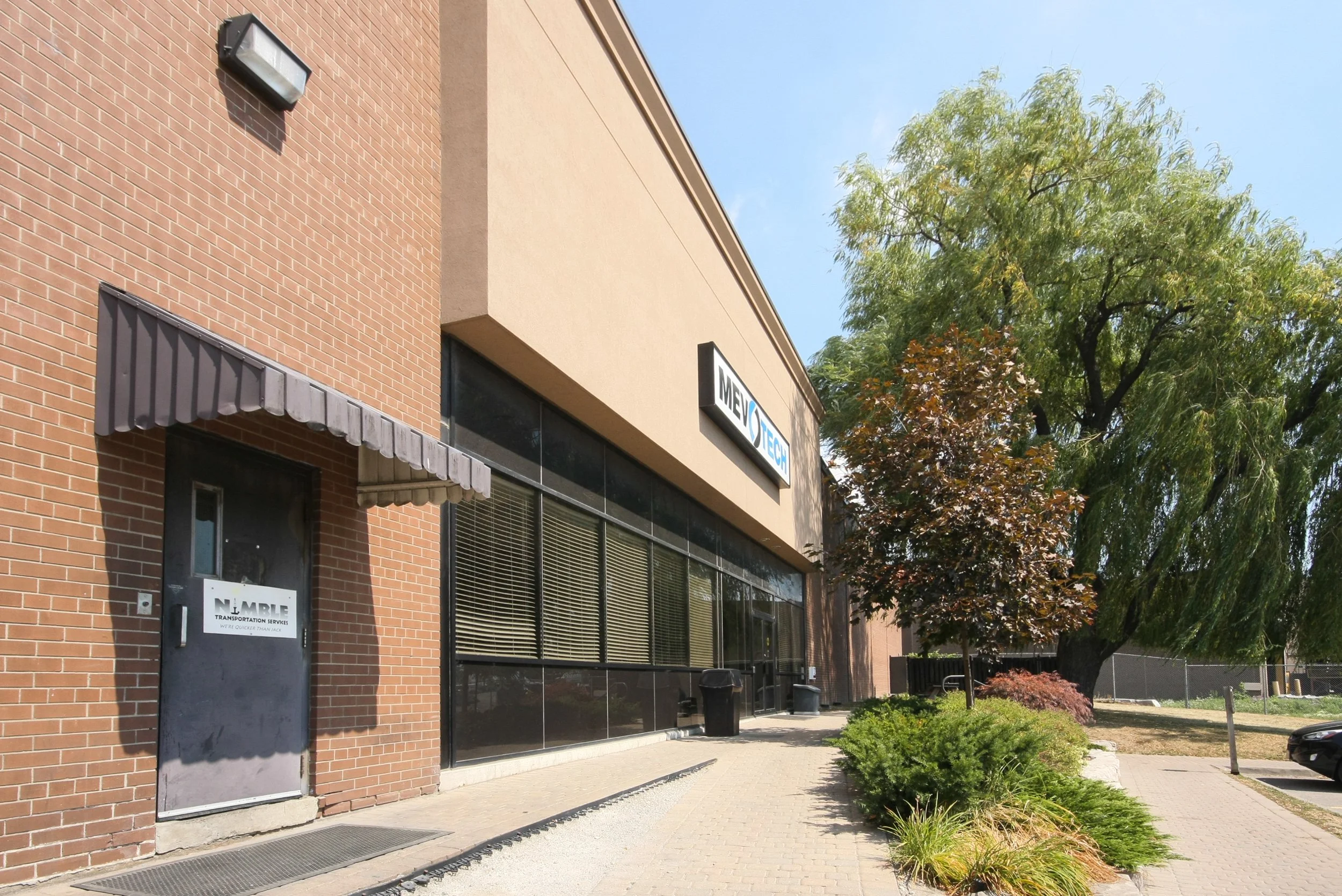 Photo of a commercial building with brick and beige walls, featuring a sign for 'MEVOTECH' and another sign for 'NPL', with trees lining the sidewalk in front and a parking lot visible on the right.
