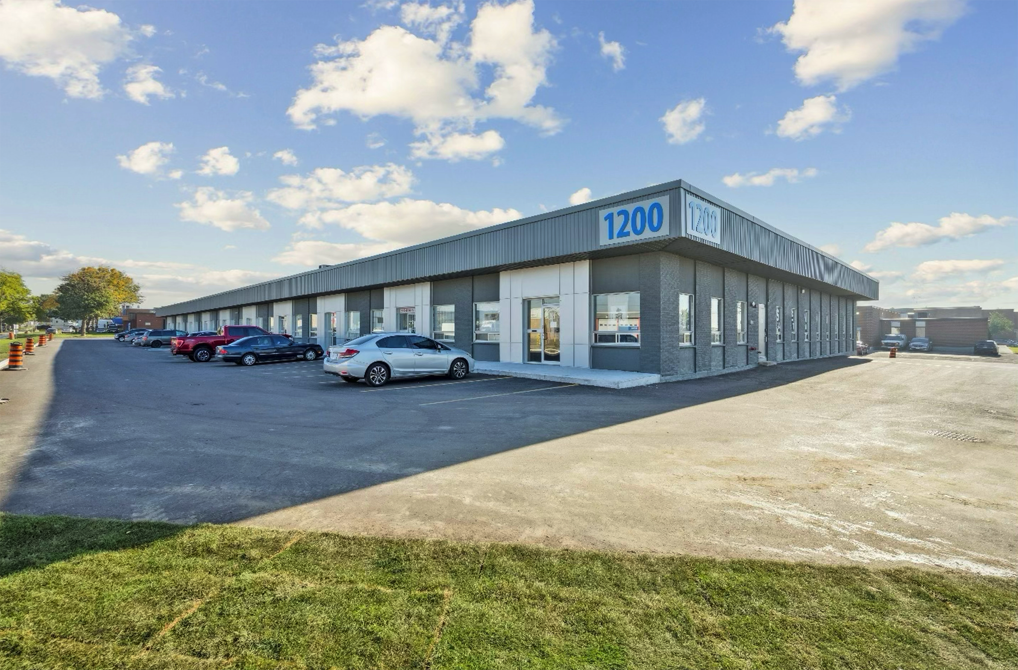 A single-story commercial building with a large blue number 1200 on the front corner, surrounded by a parking lot with several cars, under a partly cloudy sky.