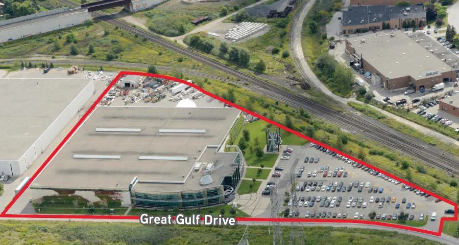 Aerial view of a commercial area outlined in red, featuring a large building with a parking lot, surrounded by greenery and nearby train tracks.