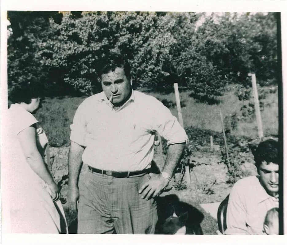 Black-and-white photo of a man with dark hair and a cigarette in his mouth, standing outdoors with trees and shrubs in the background, wearing a white shirt and checkered pants.