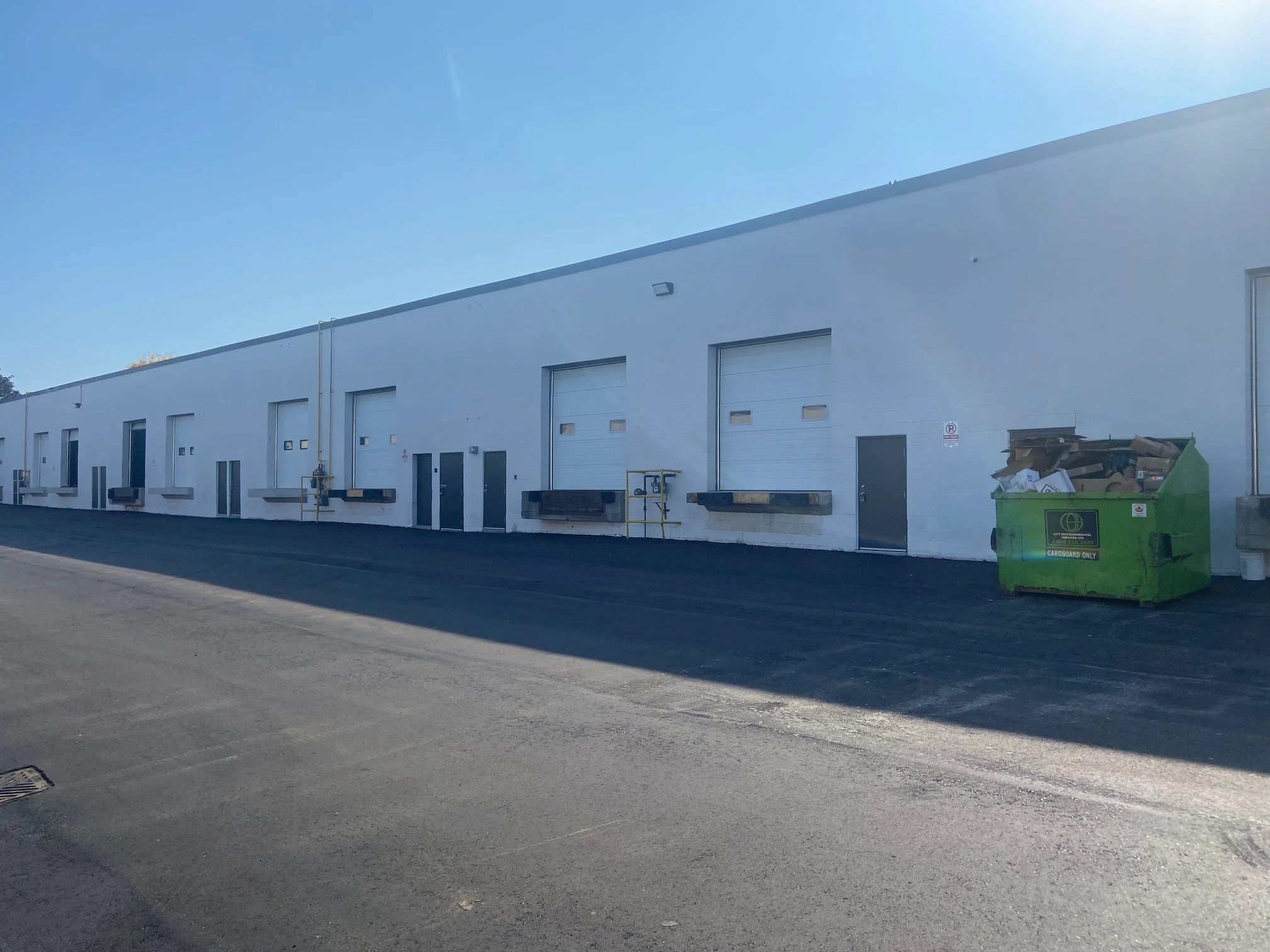 White industrial or warehouse building with multiple garage doors, a green recycling bin filled with cardboard, and a bright blue sky with sunlight glare.