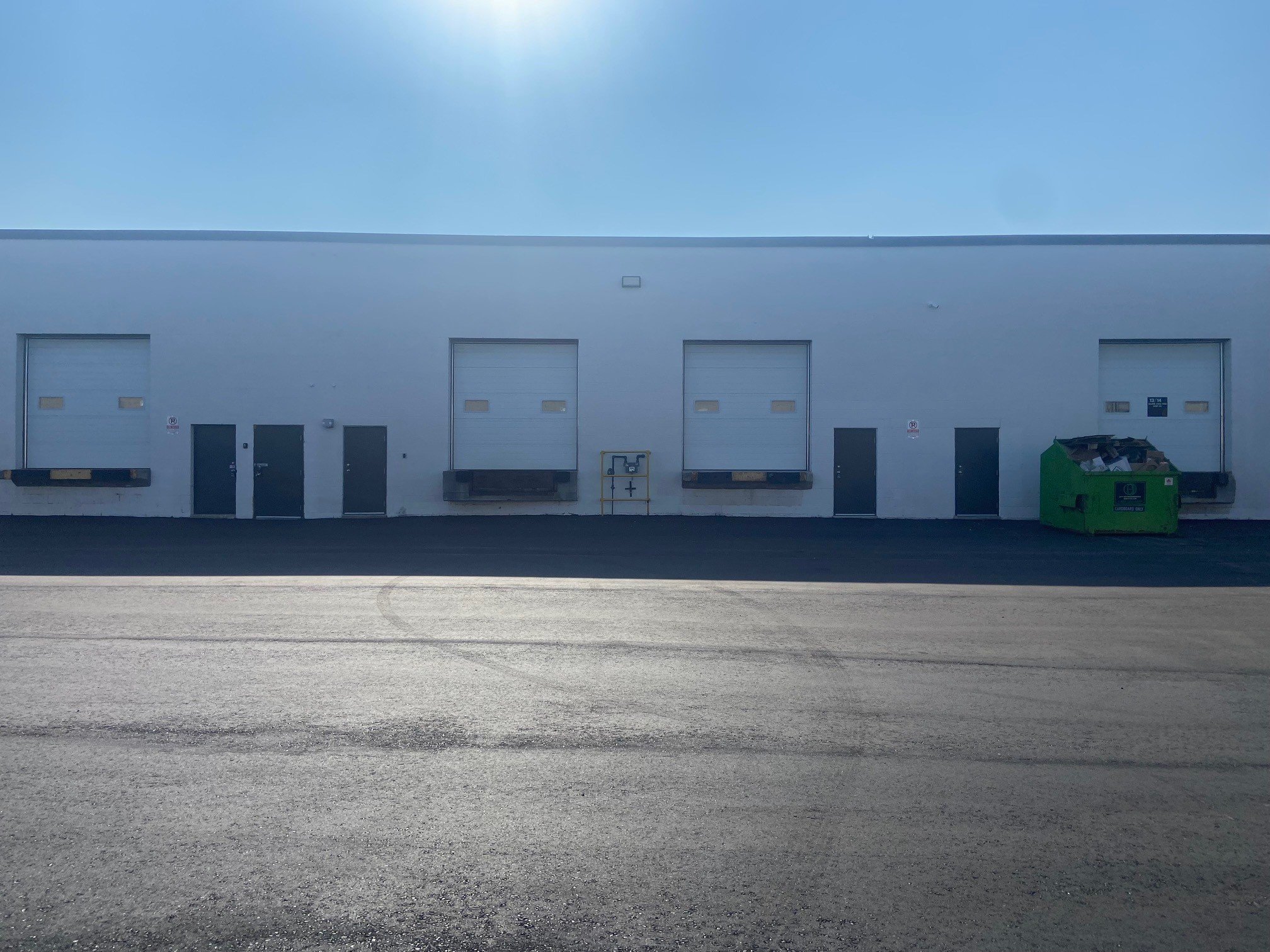 A white commercial building with three garage doors, a green dumpster on the right, and a clear blue sky with the sun shining overhead.
