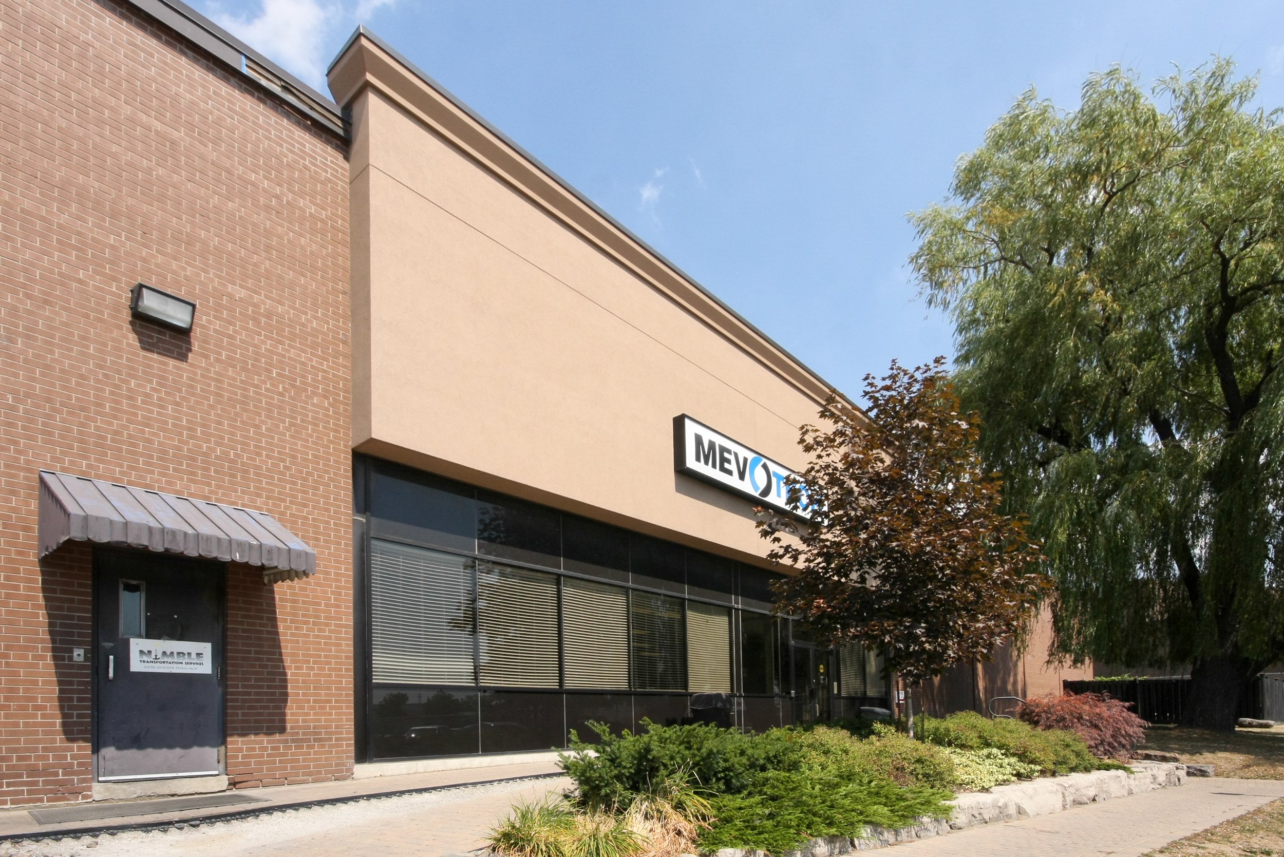 A commercial building with brick and stucco exterior, large glass windows, and a sign for MEVO, with trees and a sidewalk in front.